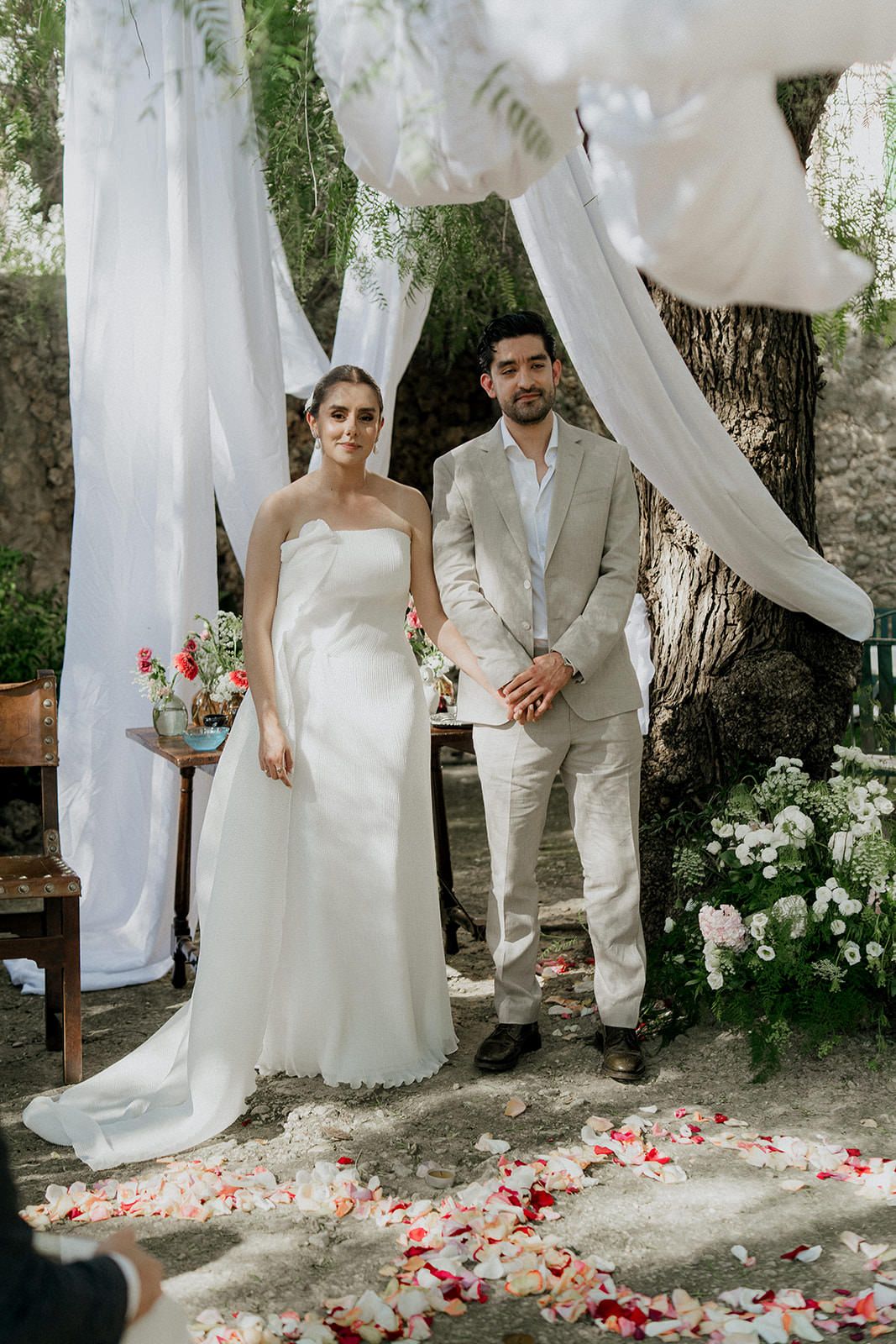 A romantic outdoor wedding scene with white flowing fabric draping, scattered rose petals, and natural garden decor.