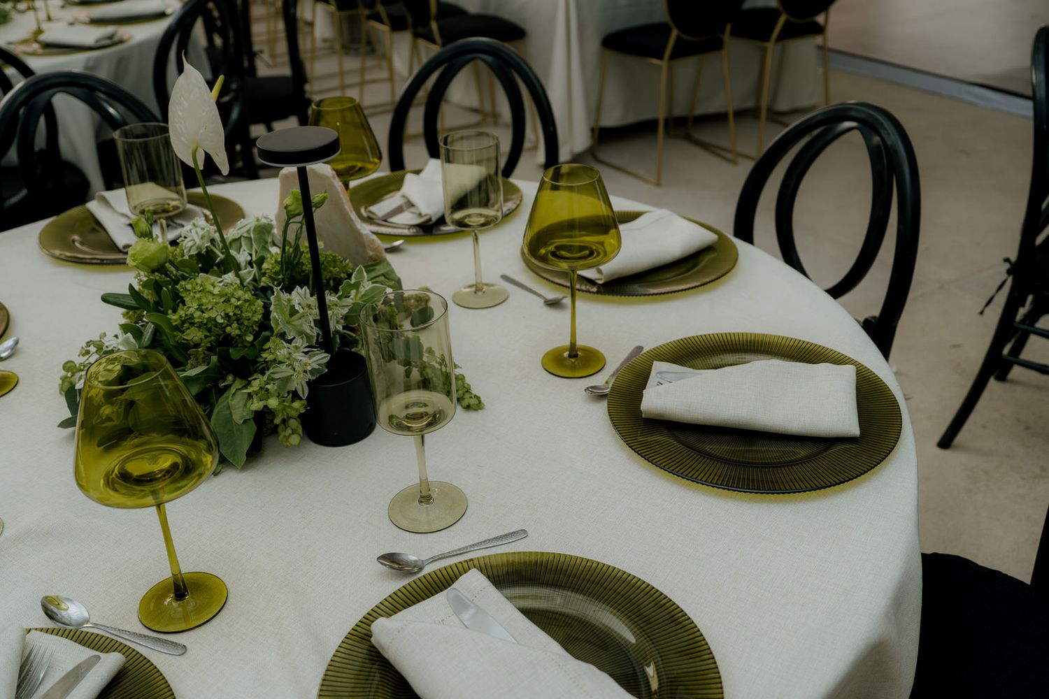 Elegant table setting with olive green chargers, wine glasses, white linens, and floral centerpiece.