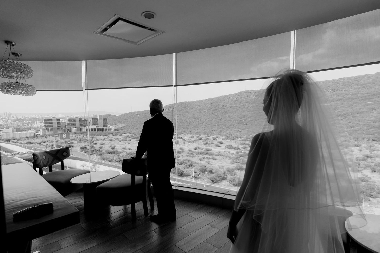 Bride and groom silhouette gazing at desert landscape through panoramic windows in black and white.