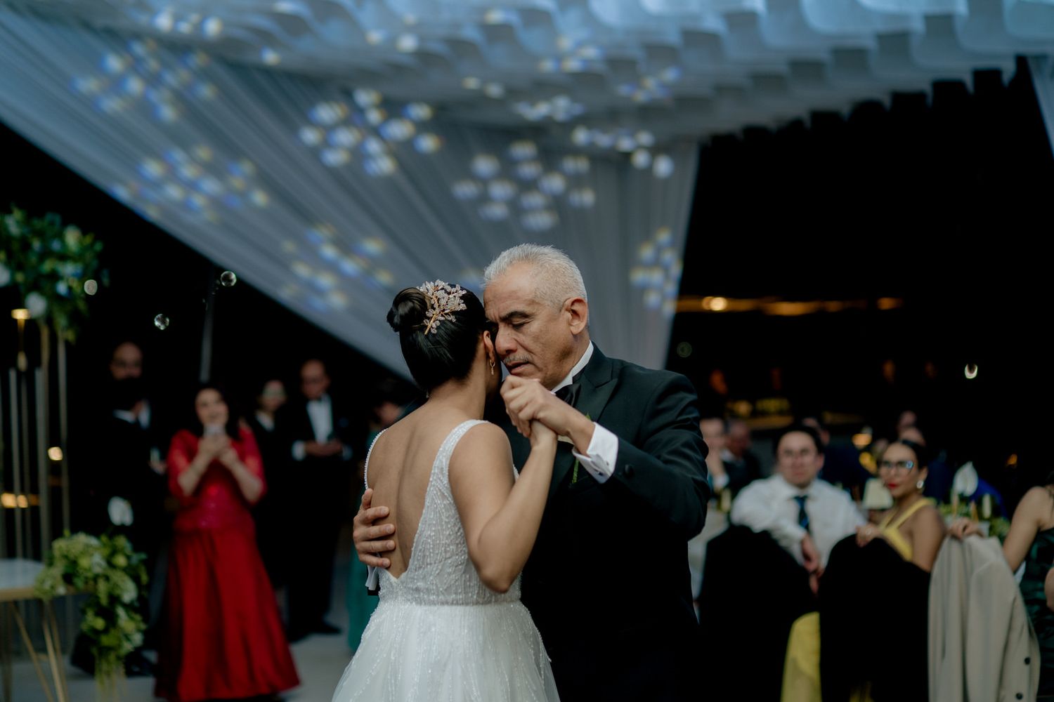 Bride and older man share emotional father-daughter first dance at elegant wedding reception with guests watching.