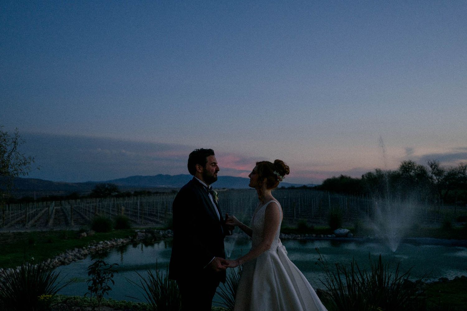 A couple stands close together during blue hour with mountains and fountains creating a romantic backdrop.