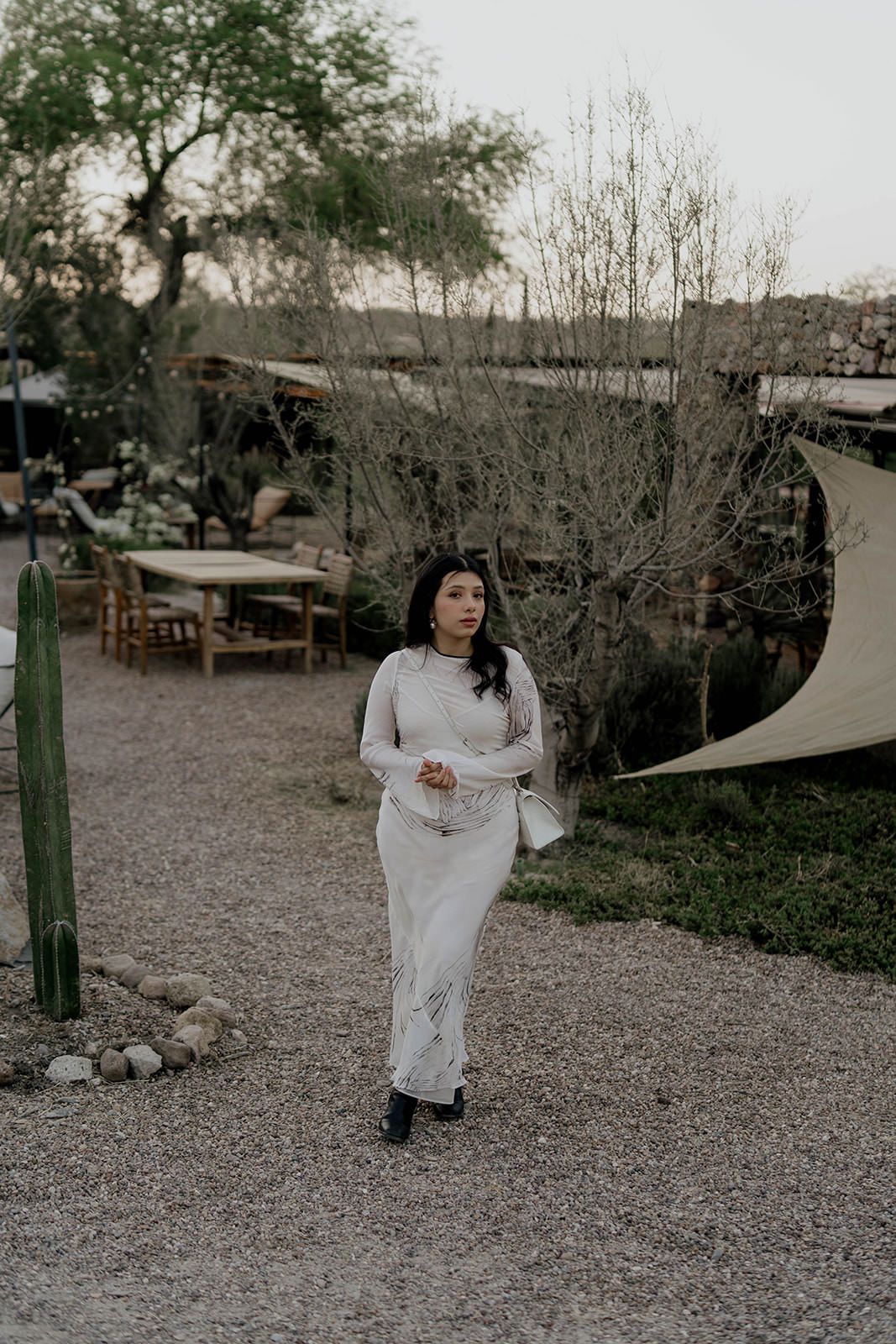 Person in flowing white dress walking on gravel path surrounded by desert landscape.