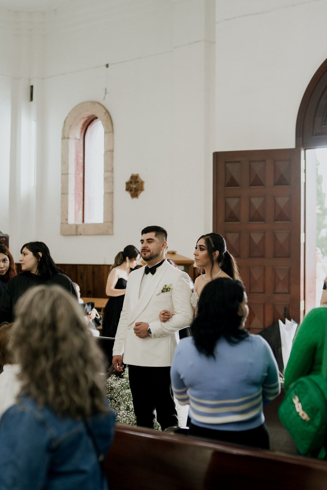 Wedding ceremony attendees seated in white church interior with wooden doors.