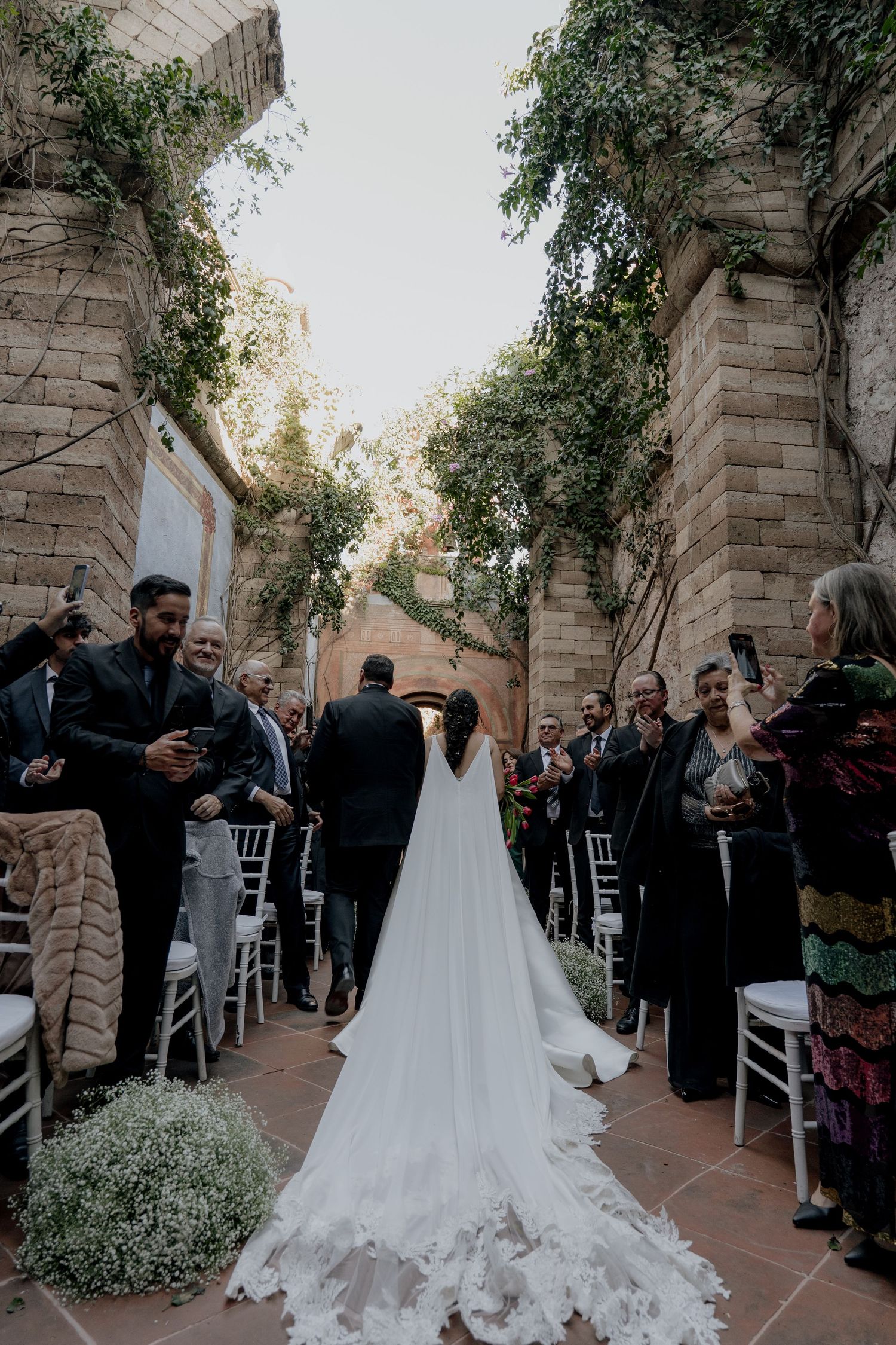 Wedding procession in an outdoor stone courtyard with trailing white dress and floral arrangements.