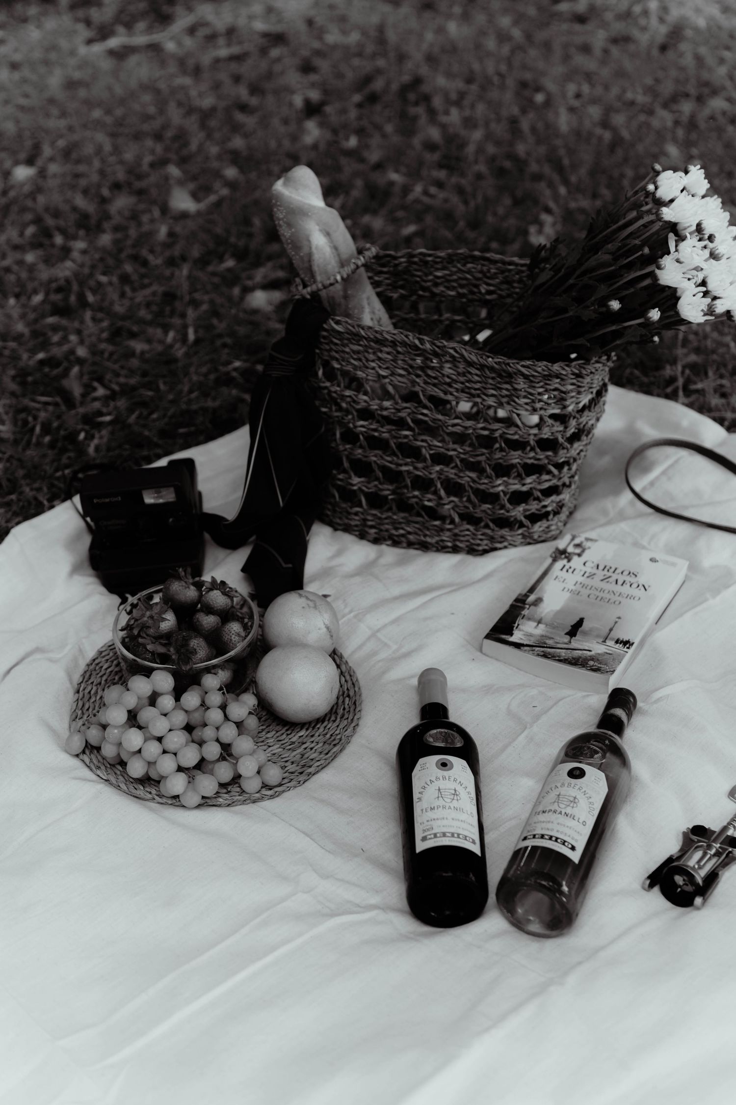 A romantic picnic setup with wine bottles, fresh fruit, and flowers on a white blanket.