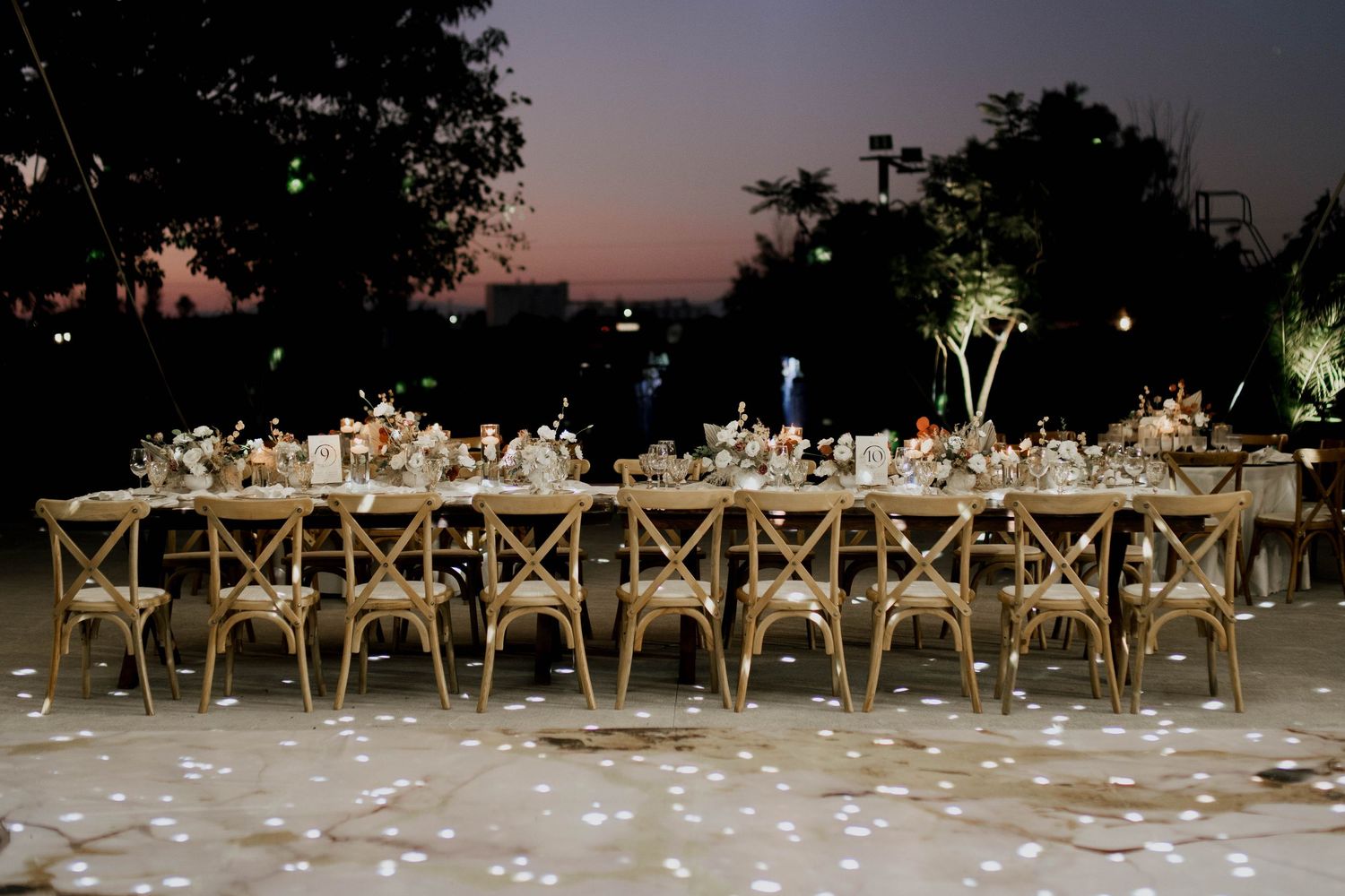 Outdoor wedding reception venue at dusk with long tables and cross-back chairs under string lights.