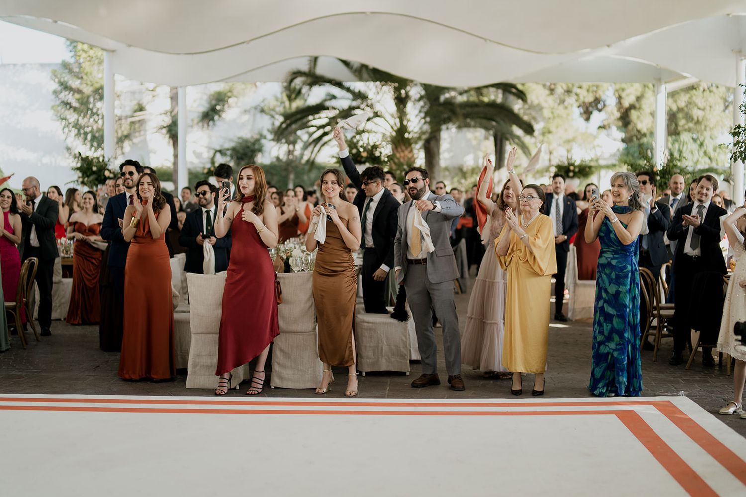Colorful wedding guests clapping and celebrating at an elegant outdoor reception under a white canopy.