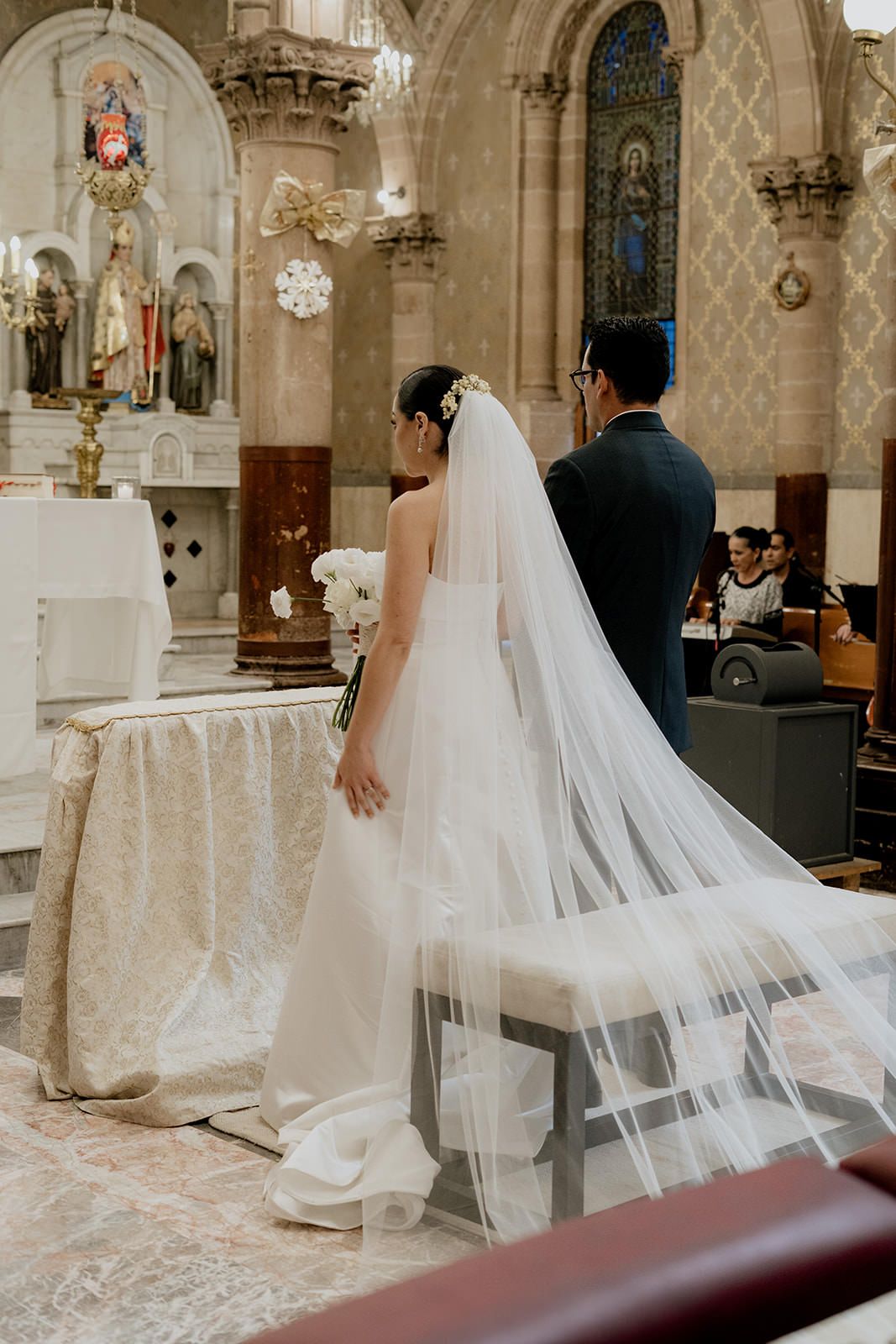 Bride in white gown with long veil stands at altar with groom during church wedding ceremony.