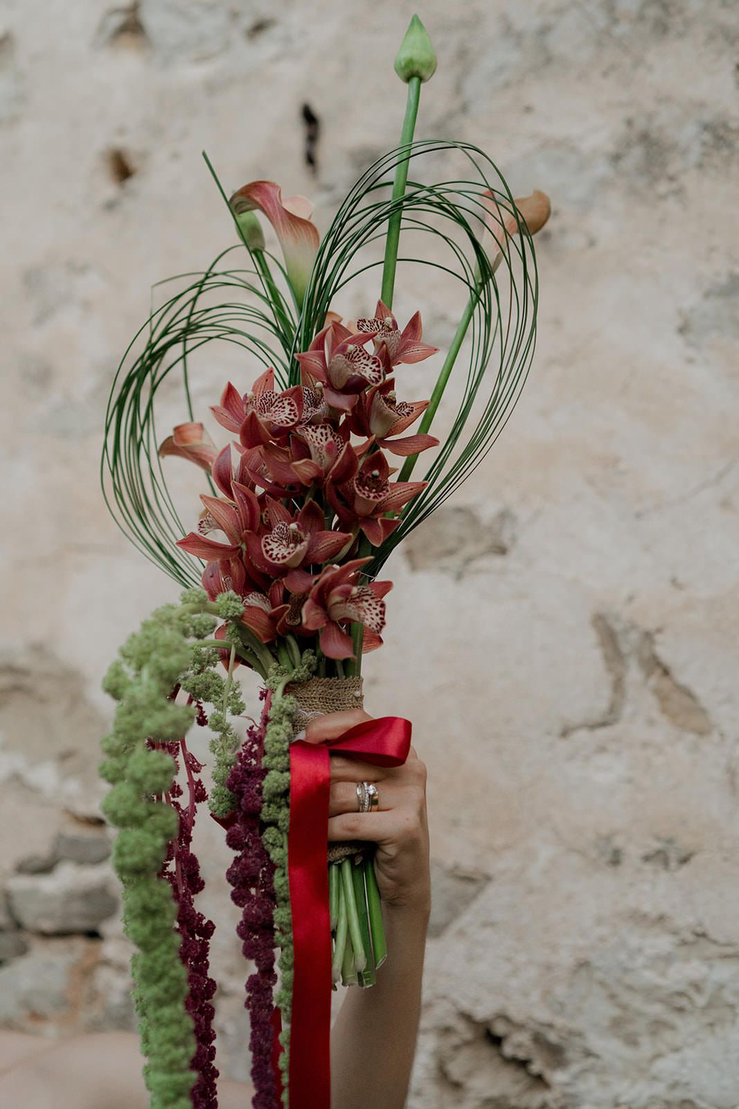 Modern floral arrangement with burgundy orchids, green amaranthus, and red ribbon against a textured stone wall.