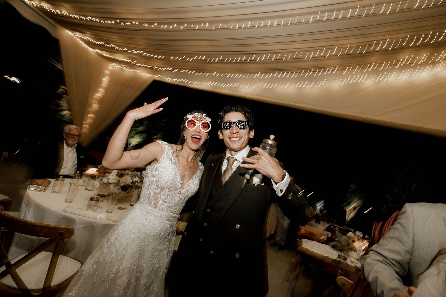 Wedding couple in sunglasses celebrate under draped fabric ceiling at reception party.