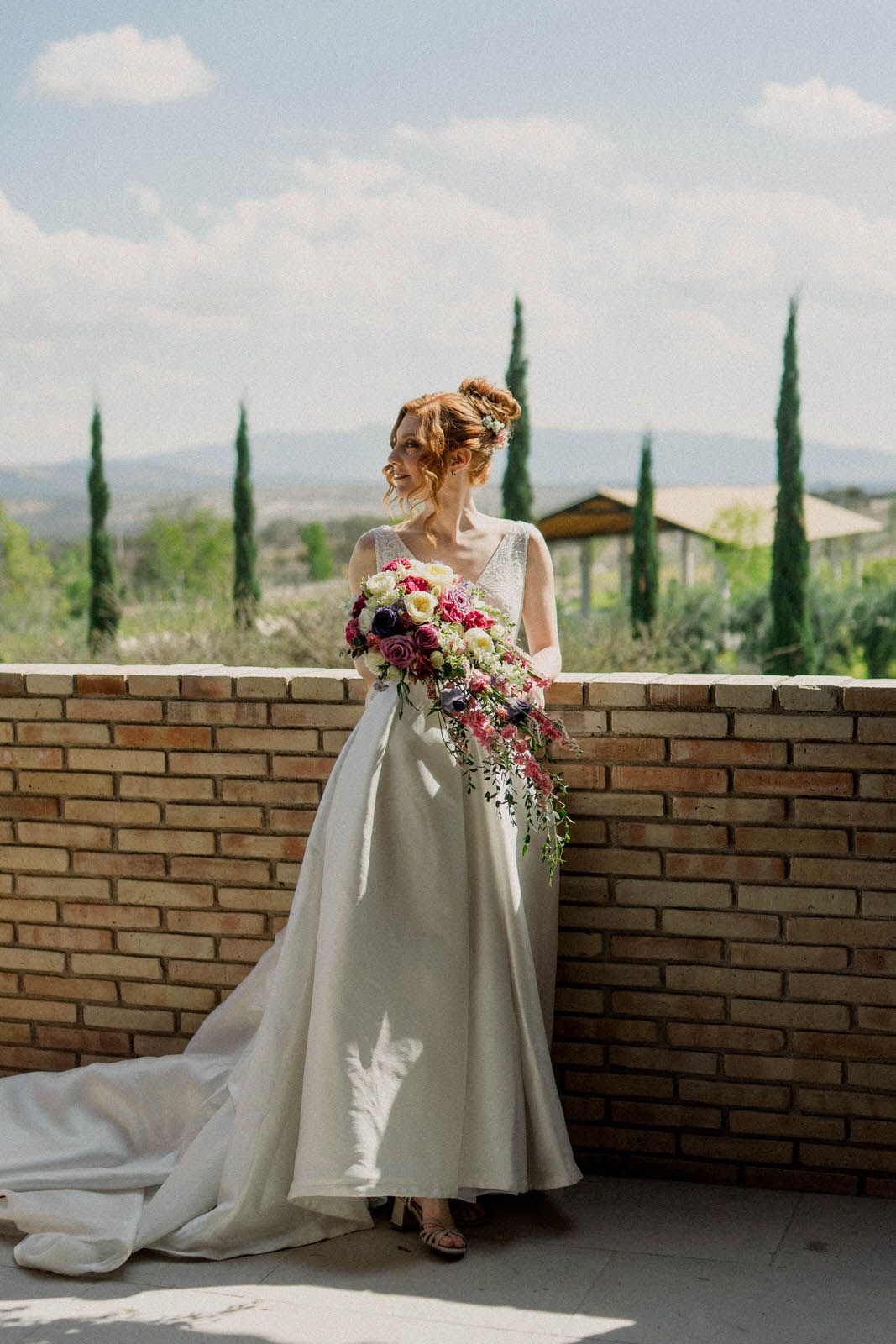 Bride in white gown poses against brick wall with Italian cypress trees in background.