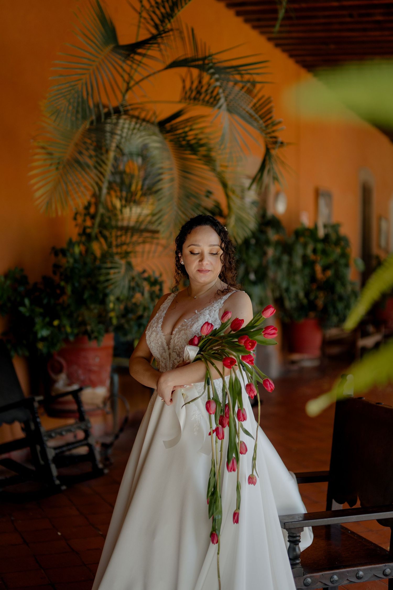 Bride in white gown holds red tulips in sunlit venue with tropical plants.