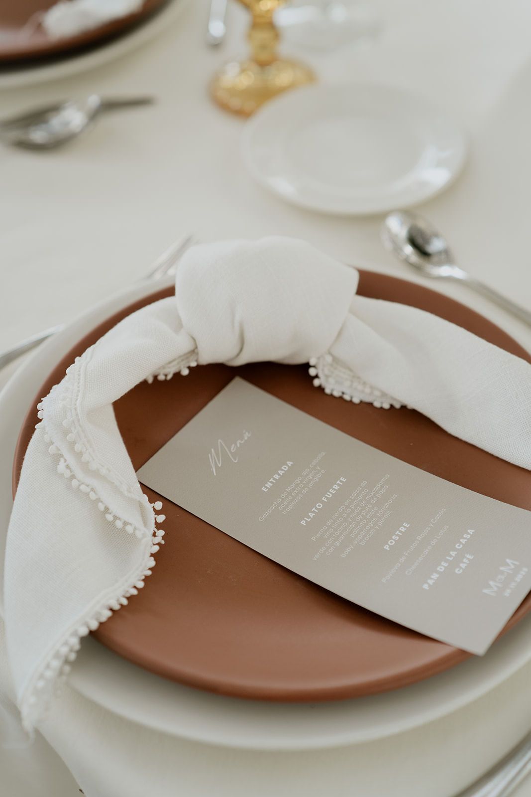 Minimalist wedding place setting with terracotta plate, white pom-pom napkin, and elegant menu card on white tablecloth.