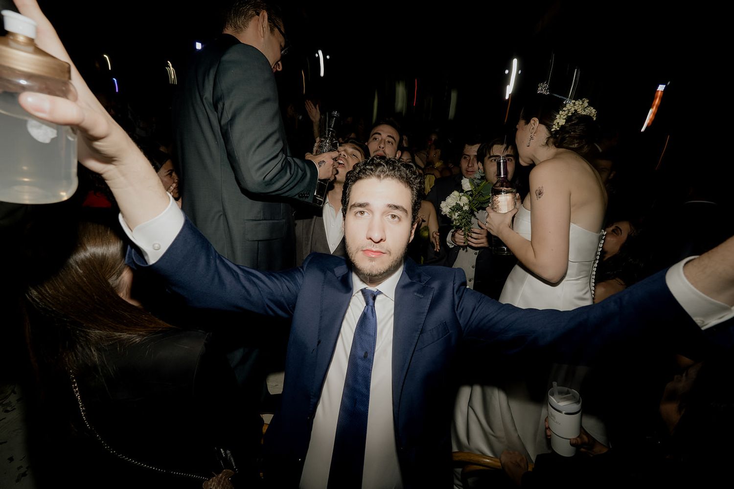 Excited groomsman in navy suit raises arms holding drinks at lively wedding reception.