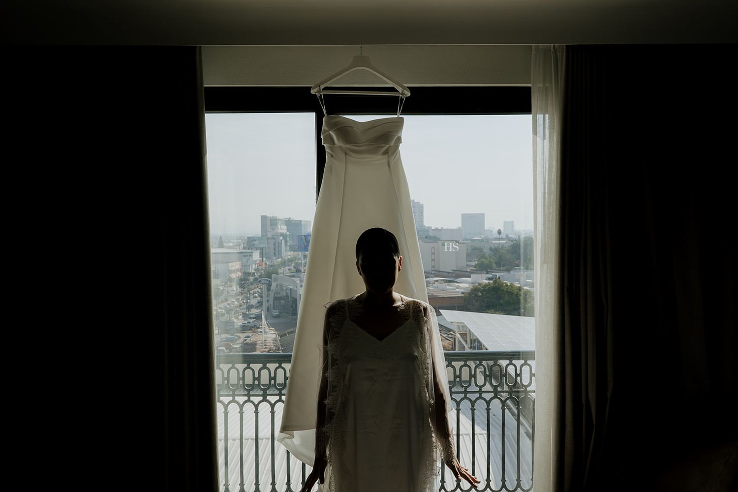 Silhouette of bride standing before hung wedding gown with city skyline view.