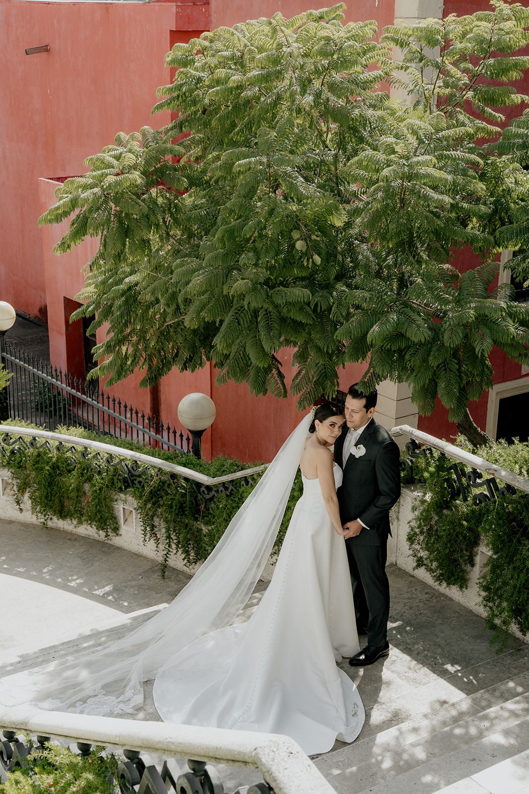 Bride and groom pose on elegant staircase with lush green trees and red building backdrop on their wedding day.