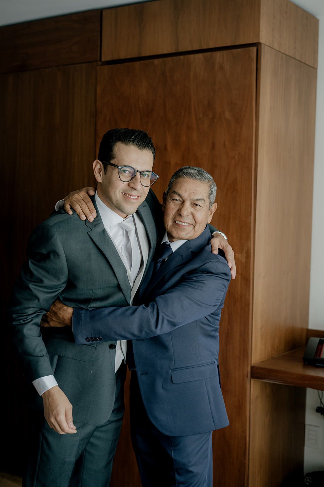Happy groom in gray suit embracing older man in blue suit in warm wedding day hug.