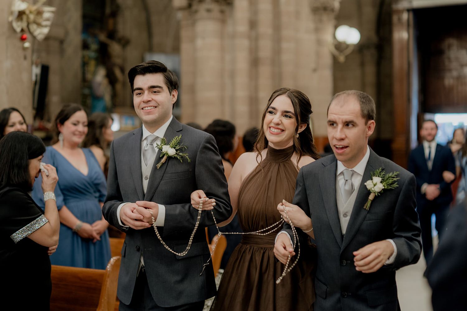 Smiling bride in brown dress flanked by two groomsmen holding decorative chain during church wedding ceremony.