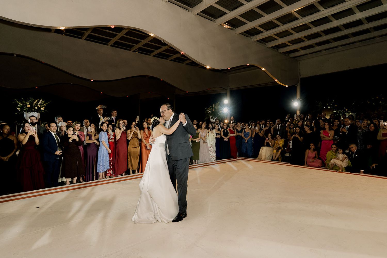 Bride and groom share first dance on white floor surrounded by wedding guests at elegant indoor reception.