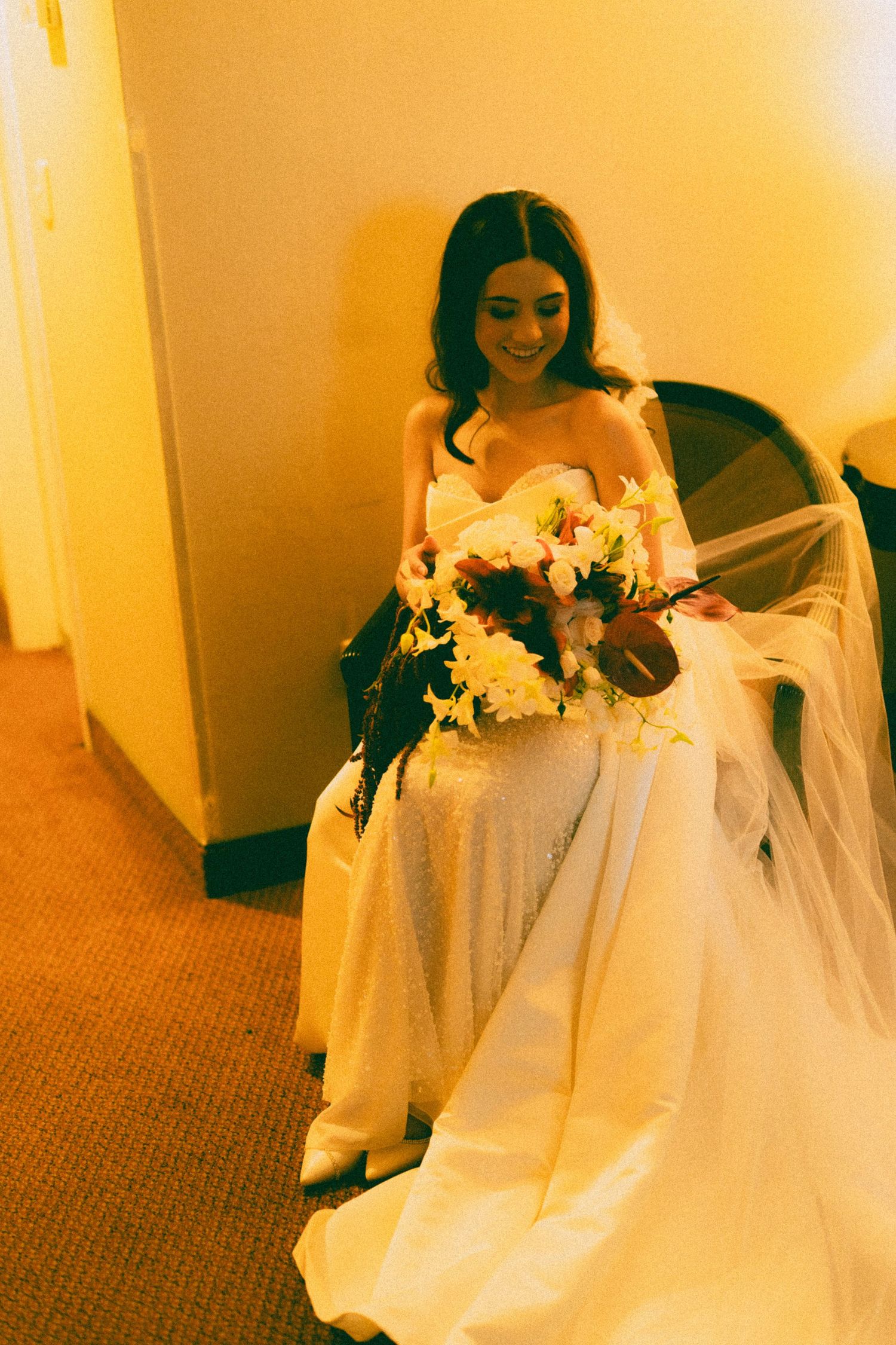 Bride in white gown holding floral bouquet, smiling while seated in warmly lit room with veil flowing.