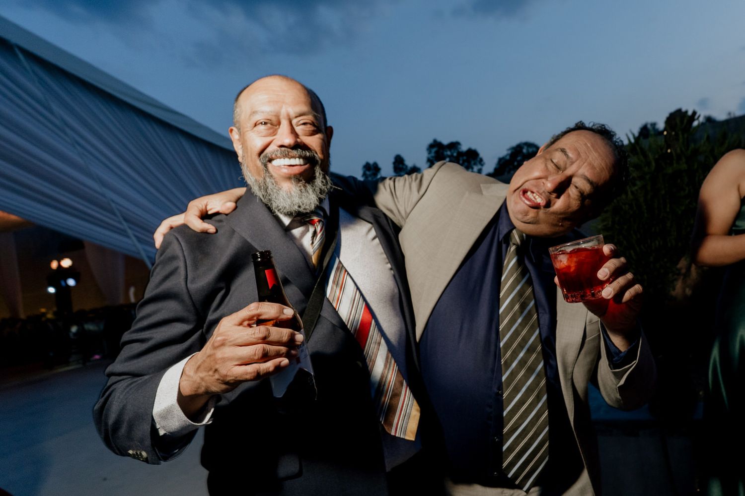 Two joyful men in suits laughing and embracing at an outdoor evening wedding reception.
