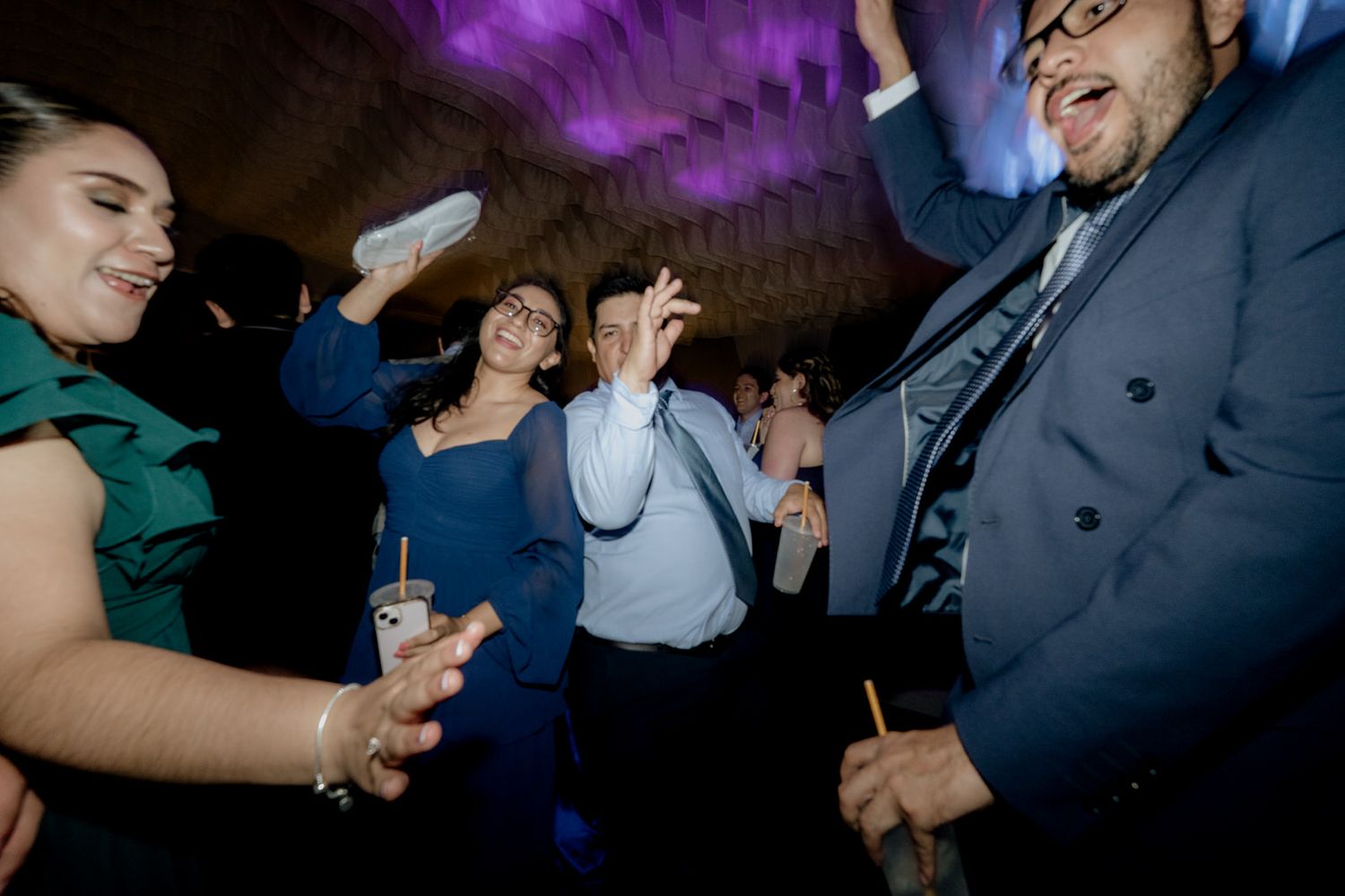 Guests dancing joyfully at a wedding reception, dressed in formal attire under purple ambient lighting.