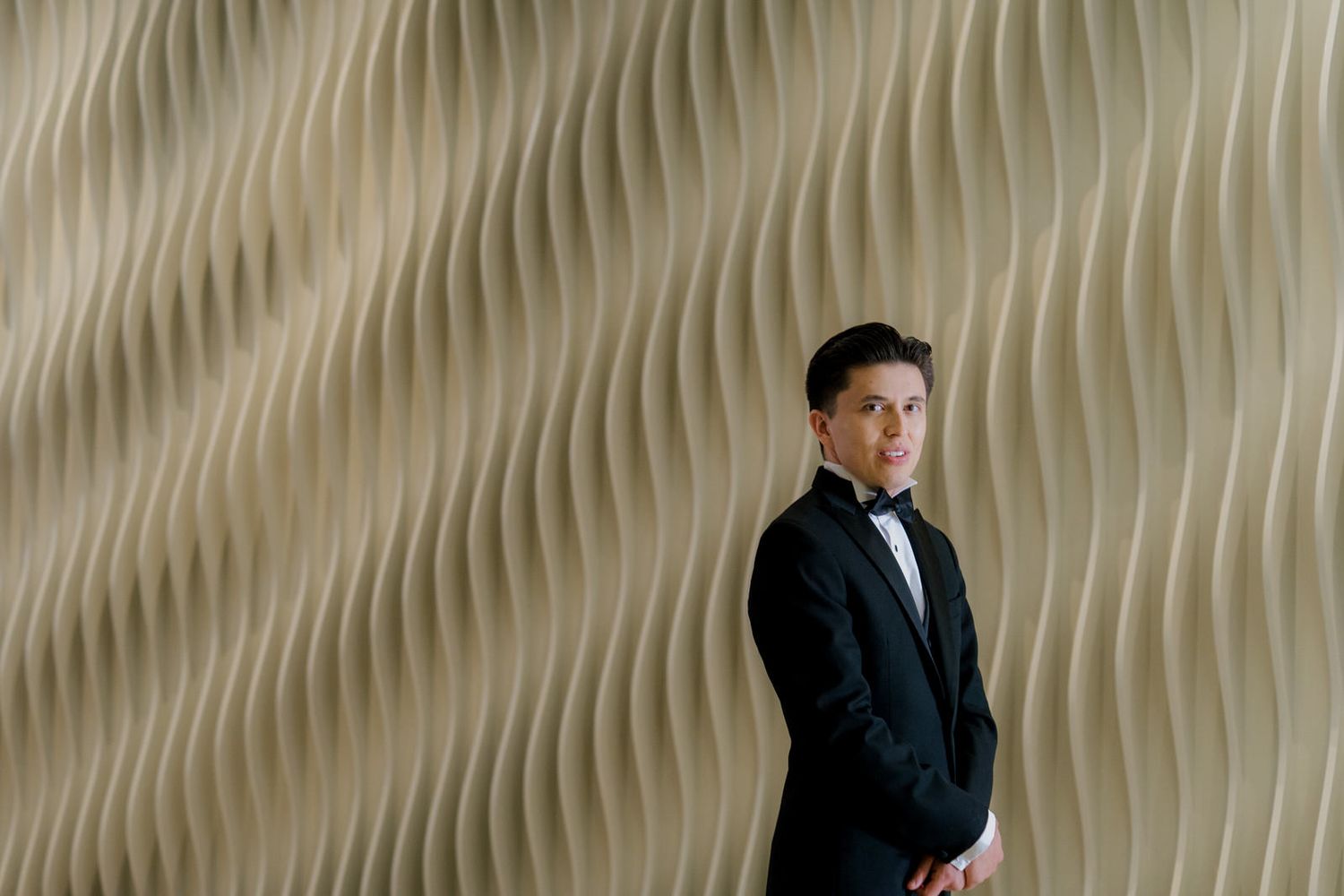 Groom in black tuxedo posing confidently against textured wavy beige wall at wedding venue.