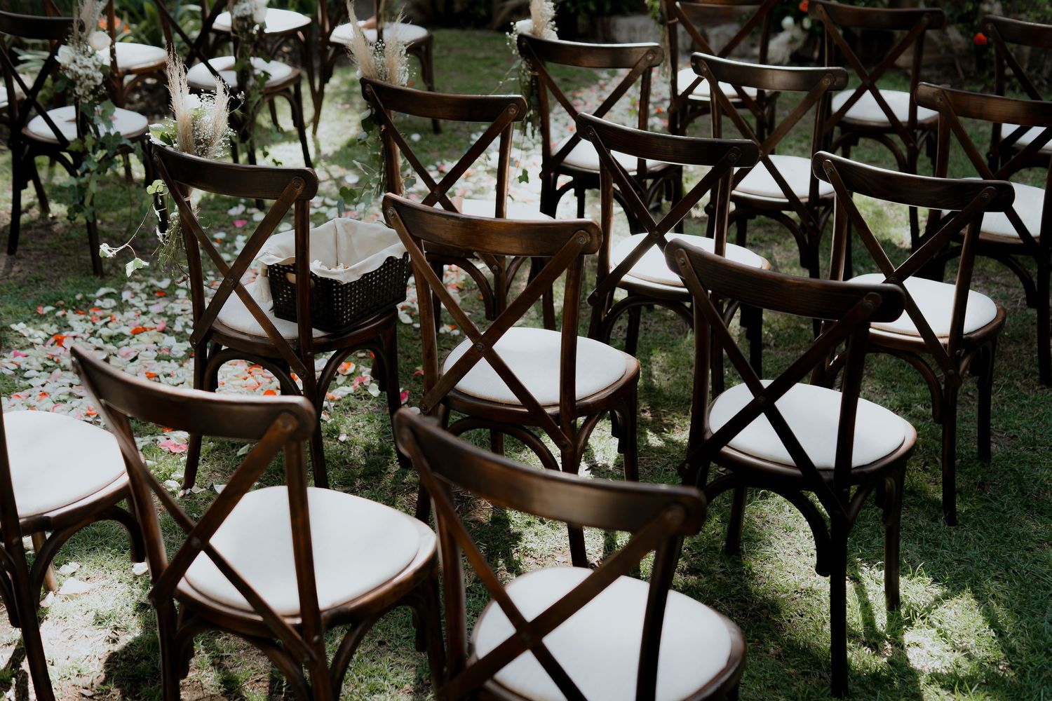 Rustic wooden crossback chairs arranged for an outdoor wedding ceremony.