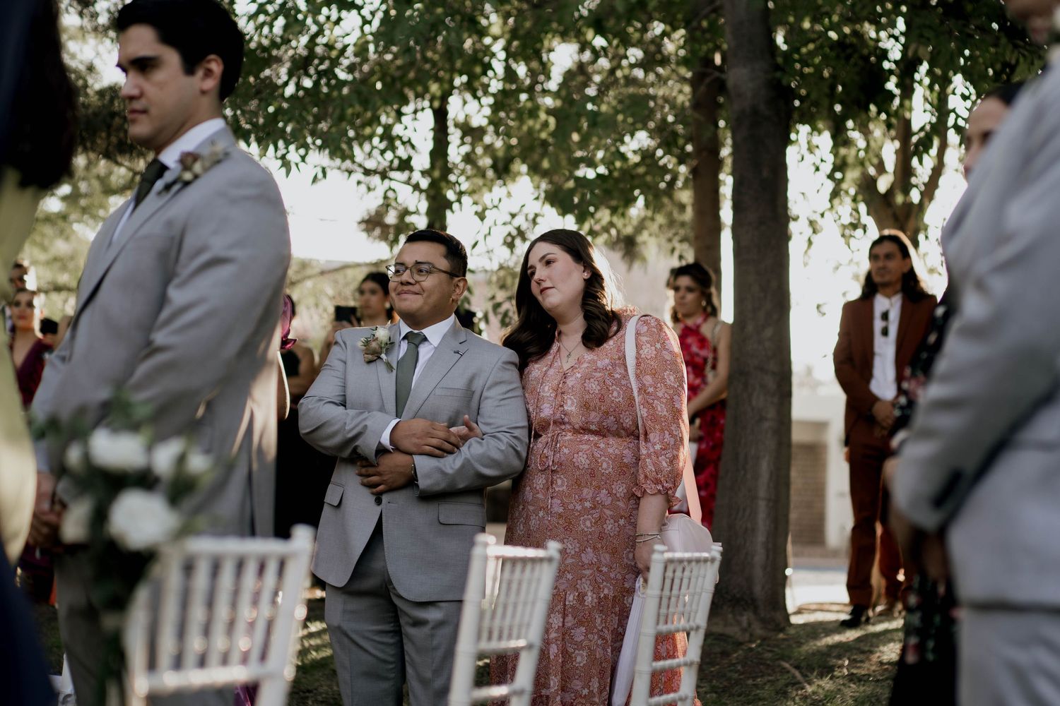 Wedding party members in grey suits and floral dresses stand during an outdoor ceremony.