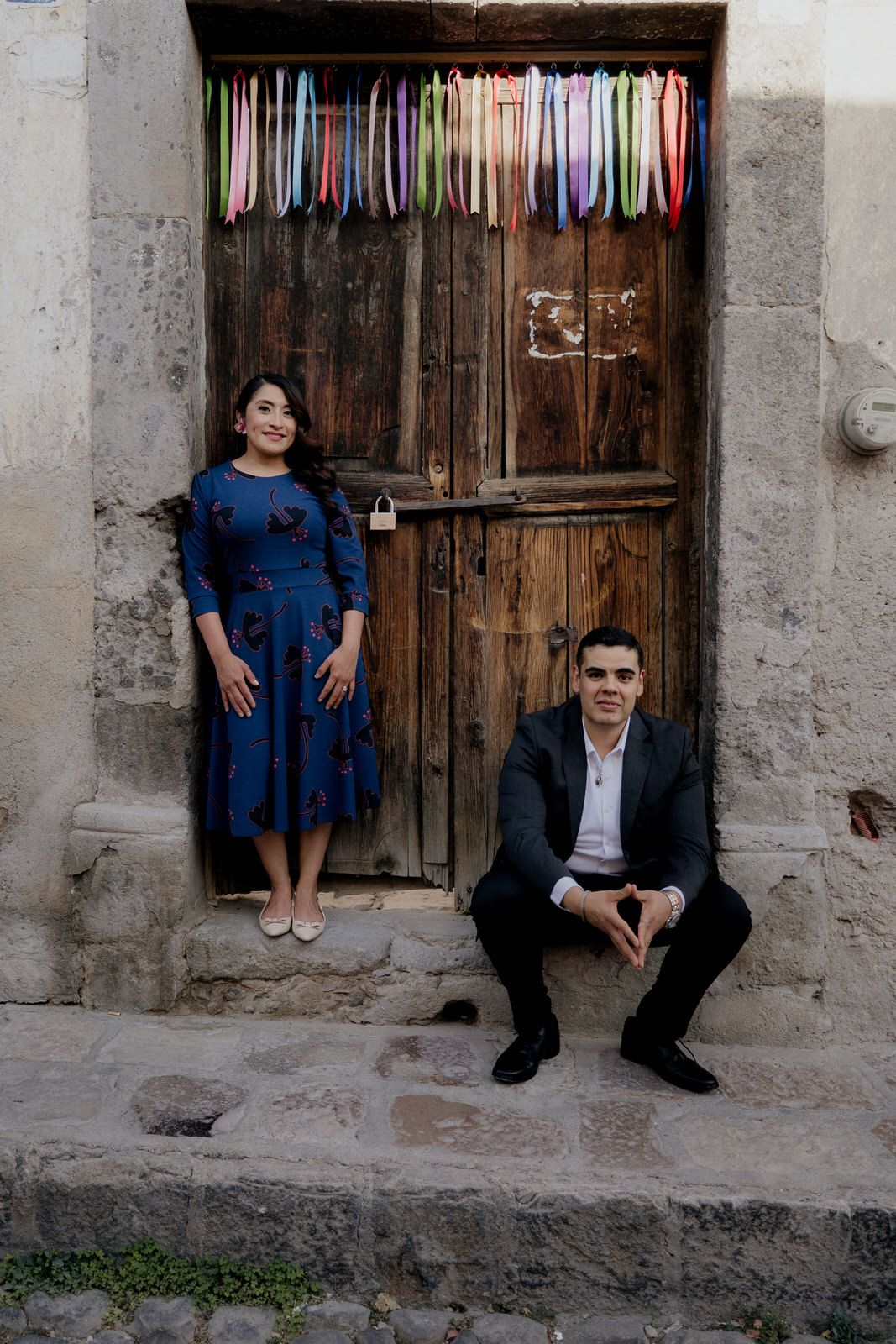 A stylish pair poses by an old wooden door decorated with colorful hanging ribbons.