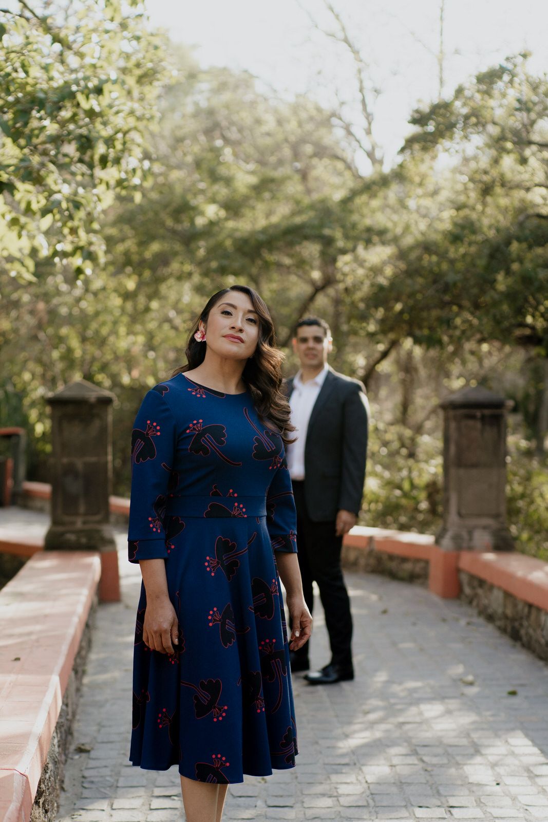 Woman in navy blue floral dress poses on stone pathway surrounded by trees.