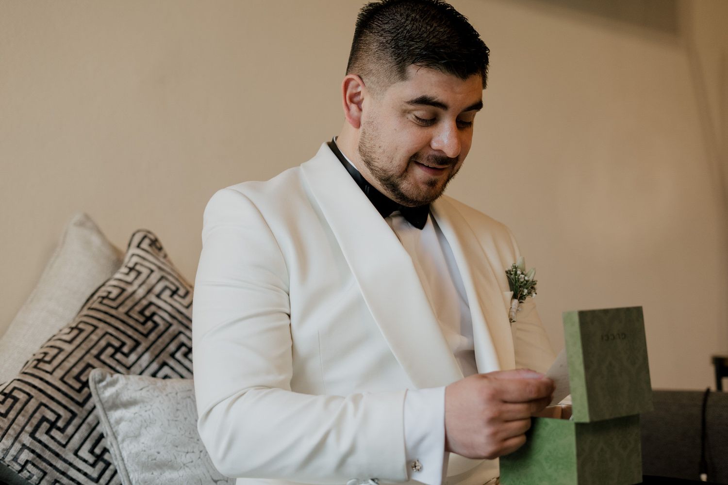 Person in crisp white tuxedo jacket examines small gift boxes while seated on couch.