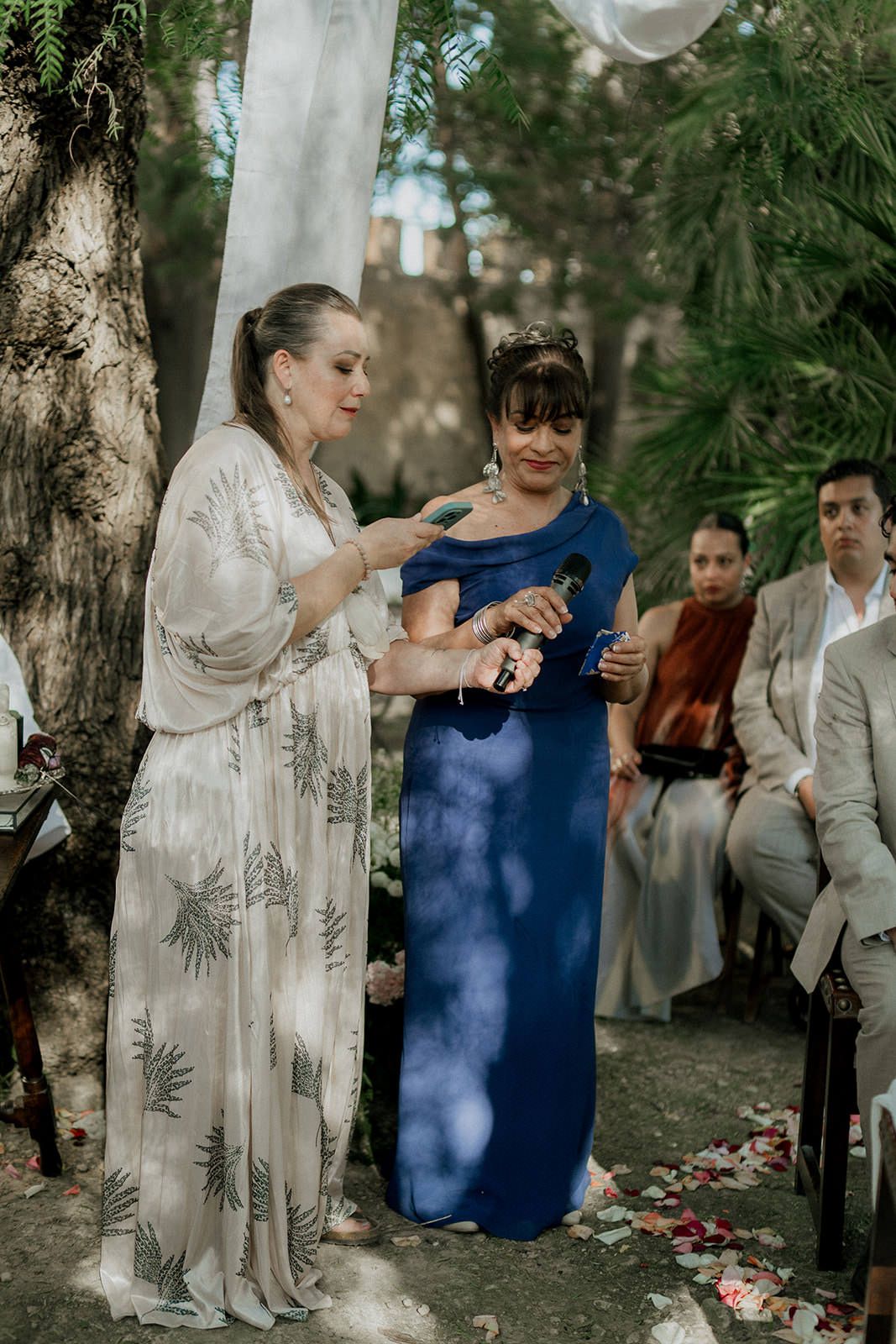 Two people share a tender moment during an outdoor wedding ceremony beneath tall trees.