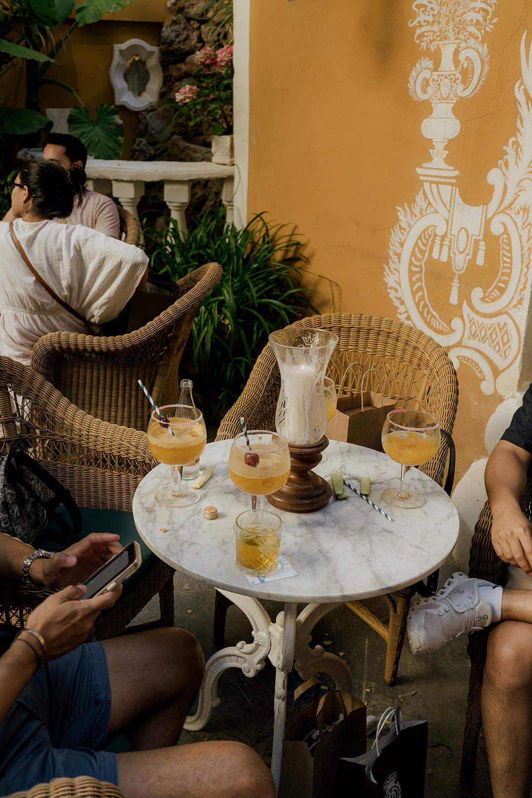 Friends enjoying cocktails and drinks at an outdoor cafe with wicker chairs and a marble table against a yellow wall with ornate decor.