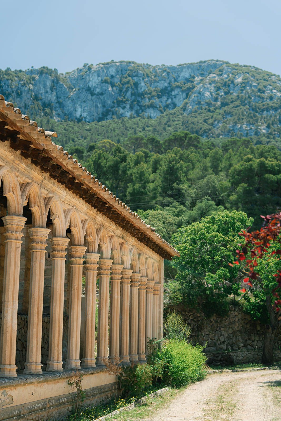 Ancient stone colonnade with arched columns stretches along a path surrounded by lush Mediterranean landscape and mountains.