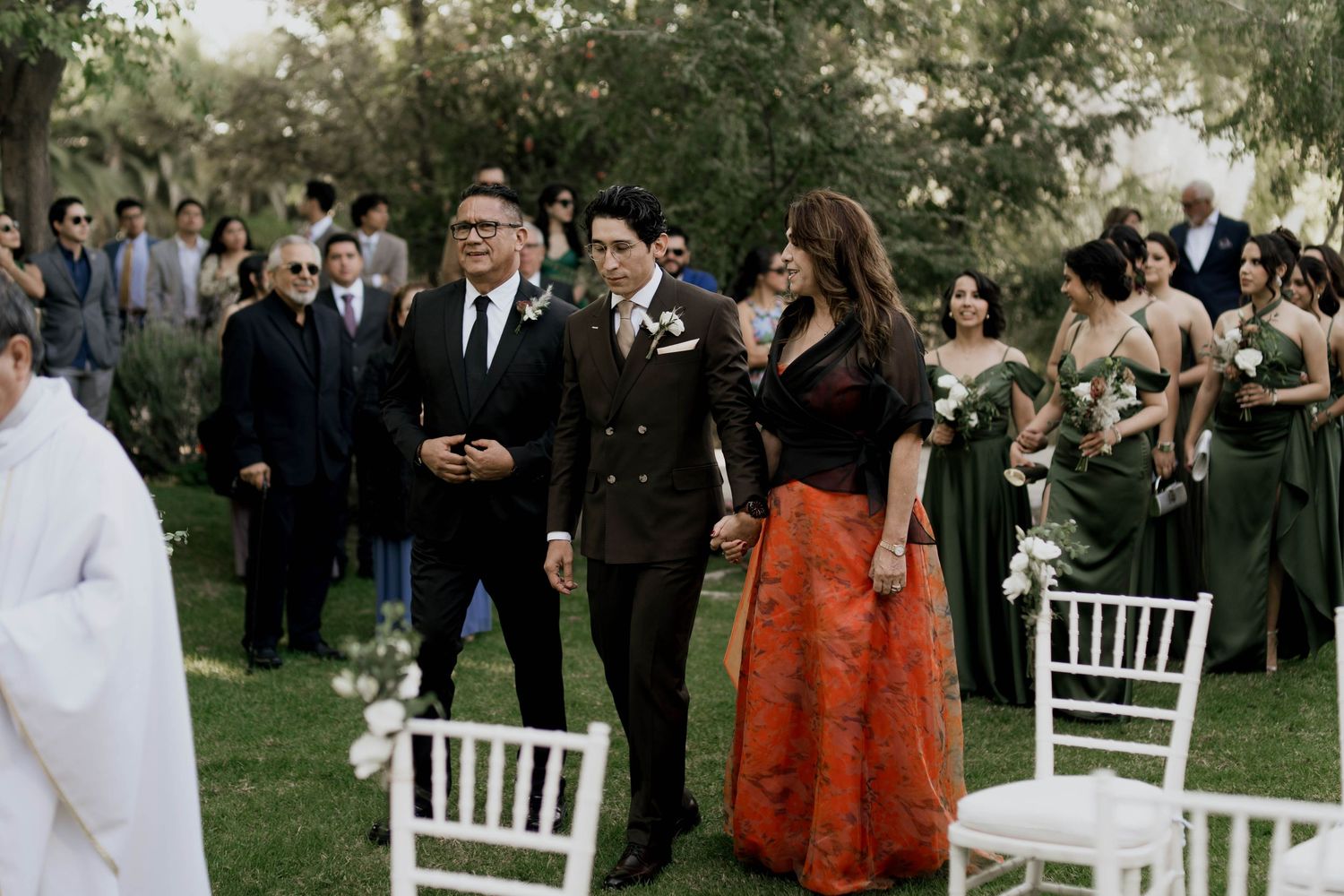 Wedding guests gather outdoors around white chairs during an elegant garden ceremony.