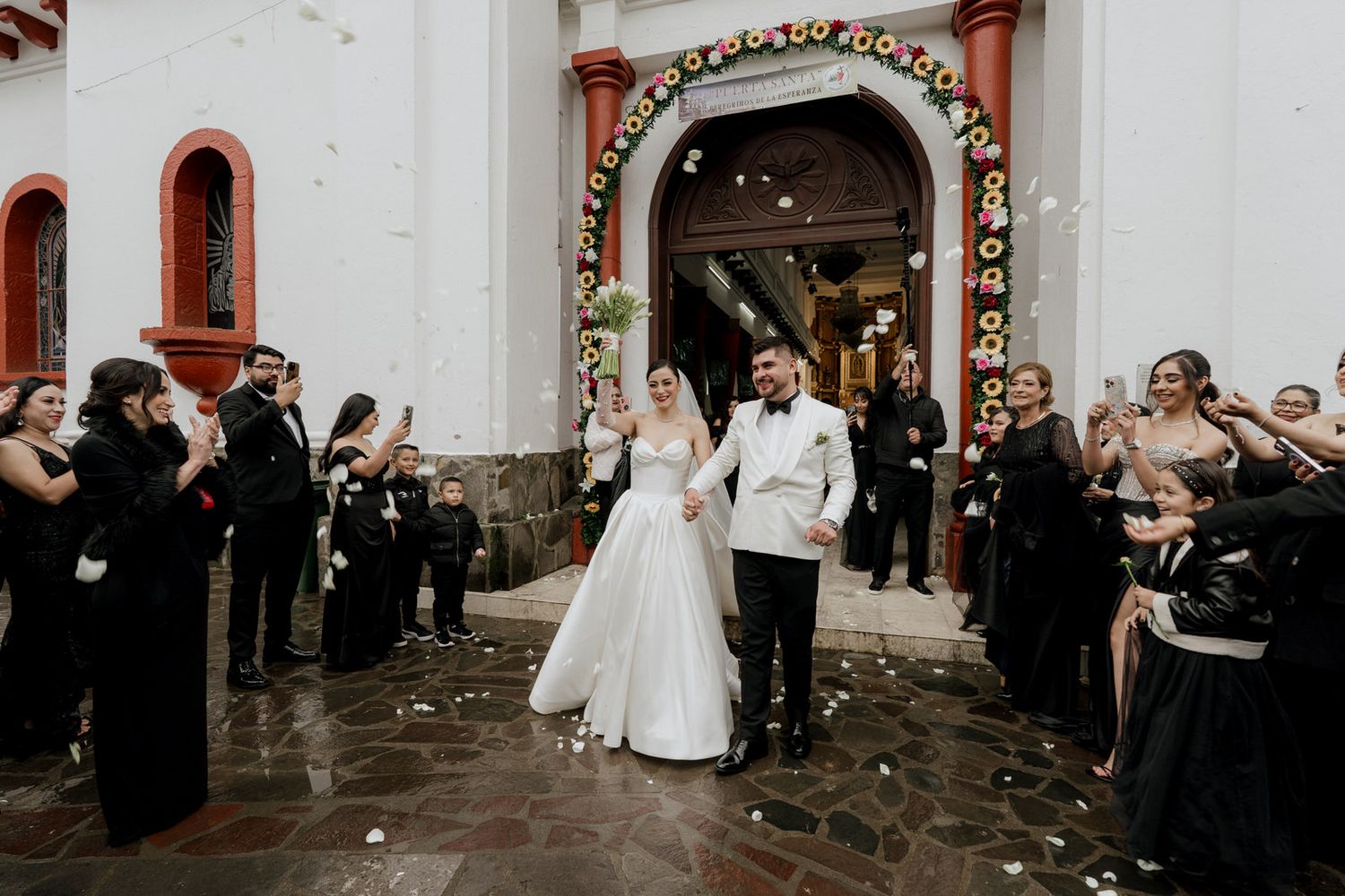 Newlyweds exit church through archway lined with guests in black attire as flower petals fall around them.