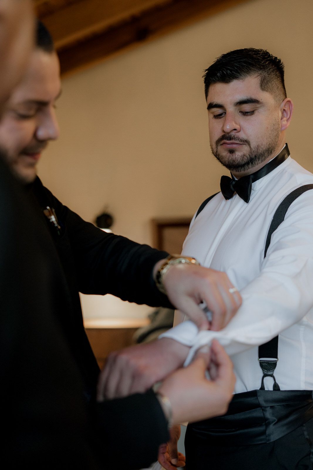 Groomsmen help adjust each other's formal attire before a wedding ceremony in a warmly lit room.
