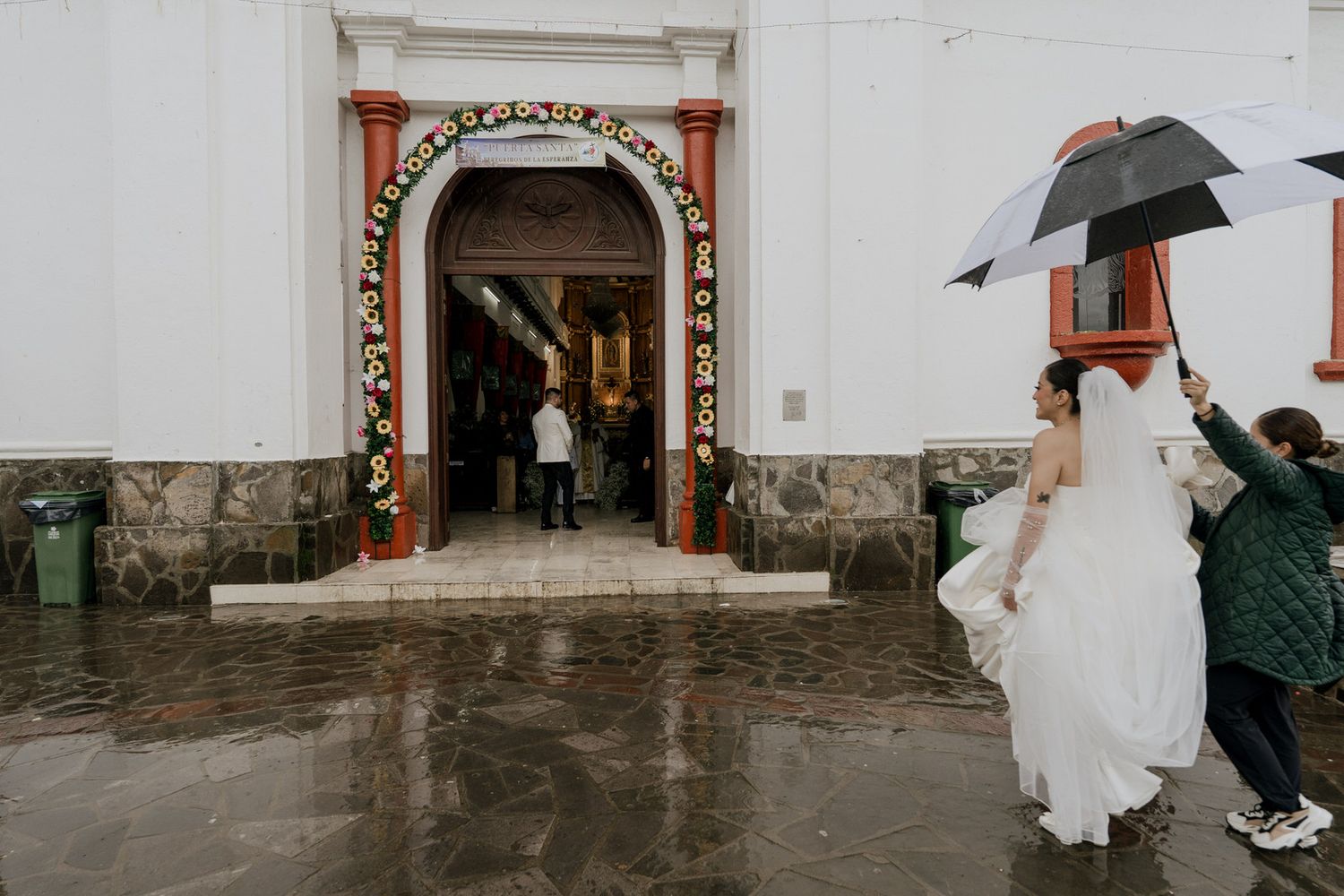 Wedding guests gather outside a white church entrance with umbrellas on a rainy day.