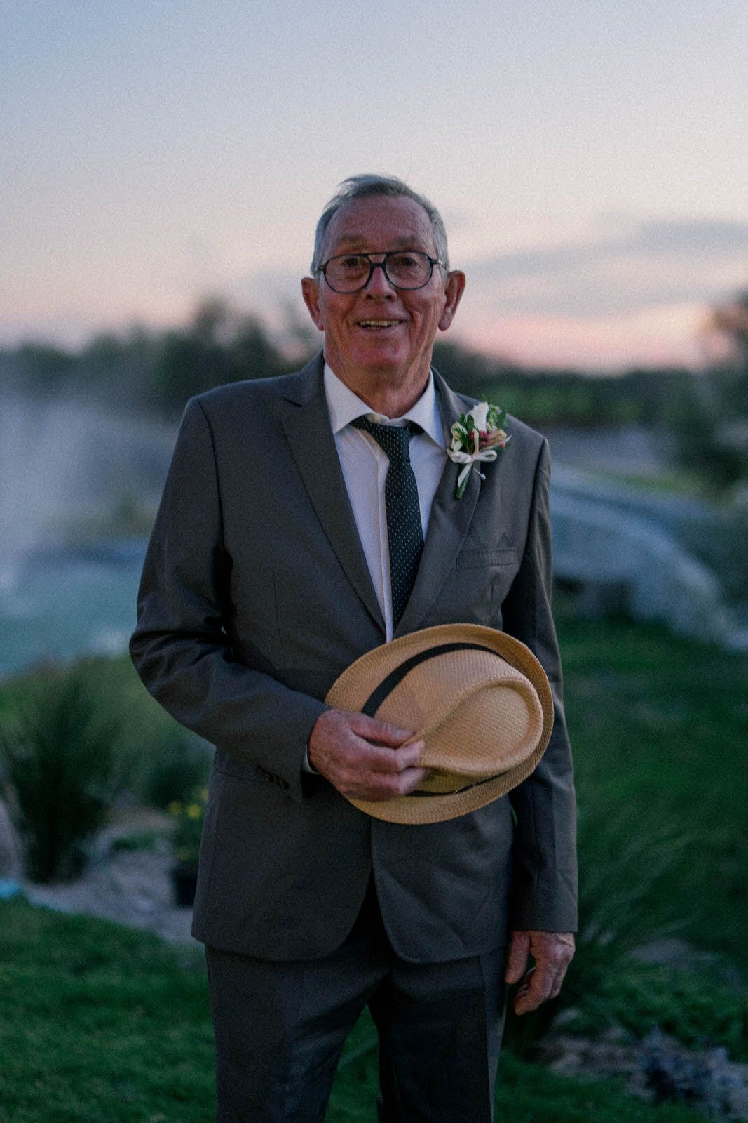An elegant gentleman in a grey suit holds a hat while standing outdoors at dusk.