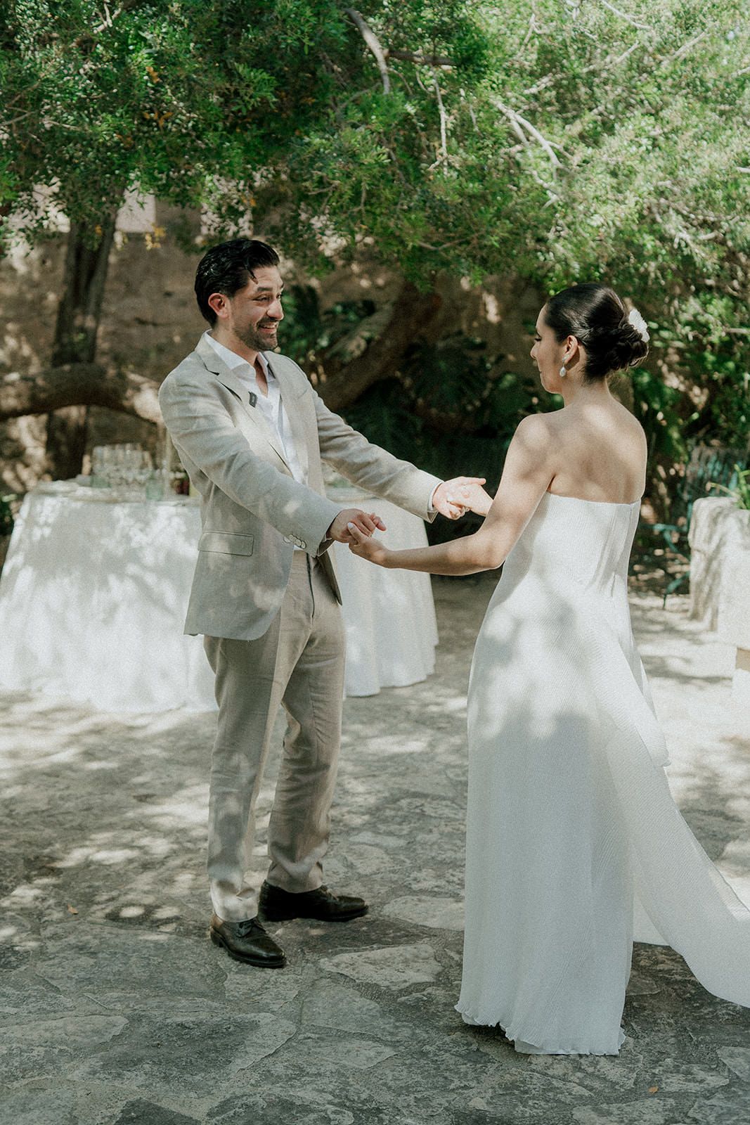 A newlywed couple twirls and dances together in a romantic garden setting with dappled sunlight.