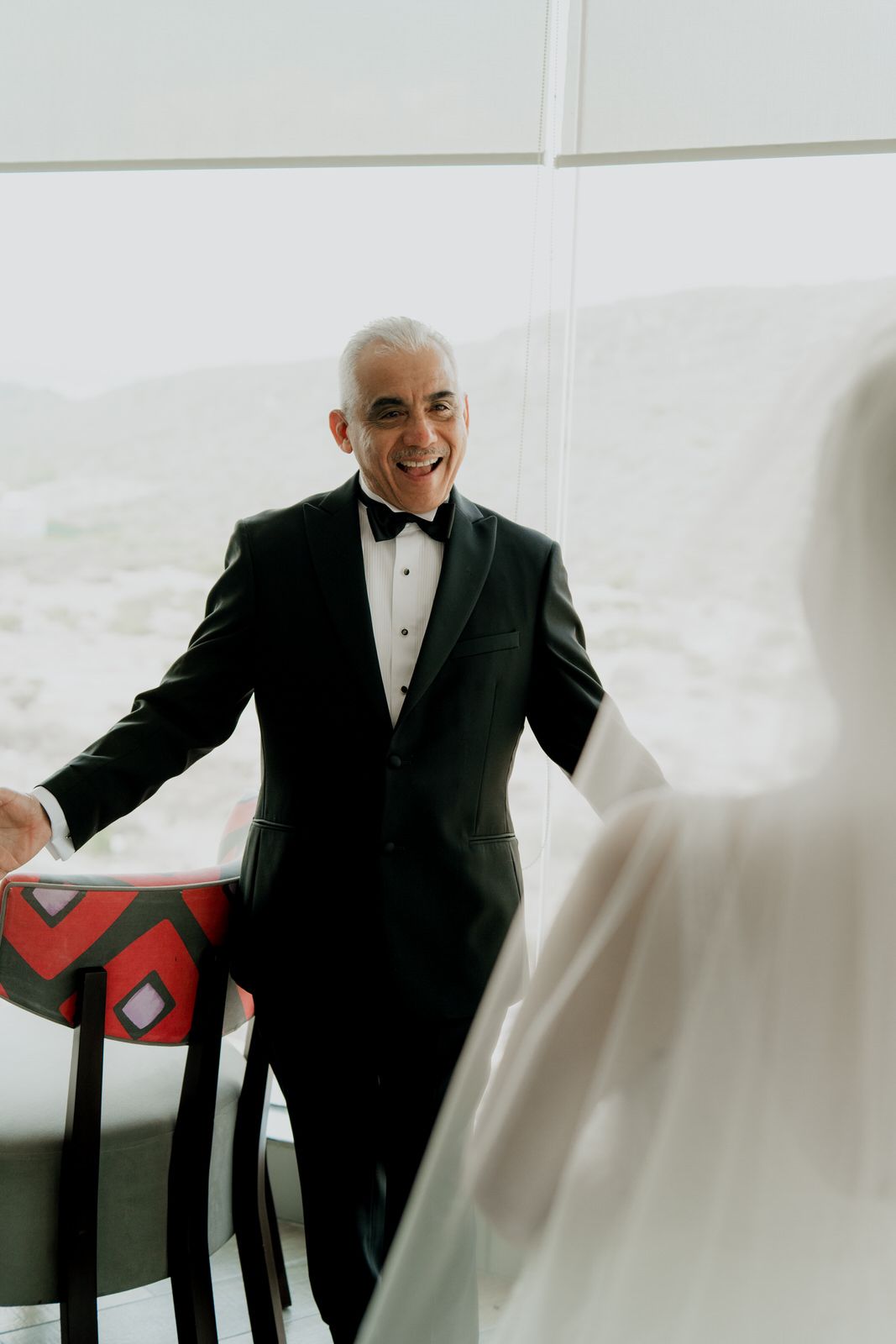 Smiling groom in black tuxedo and bow tie reacts joyfully to bride in white veil during first look wedding moment.
