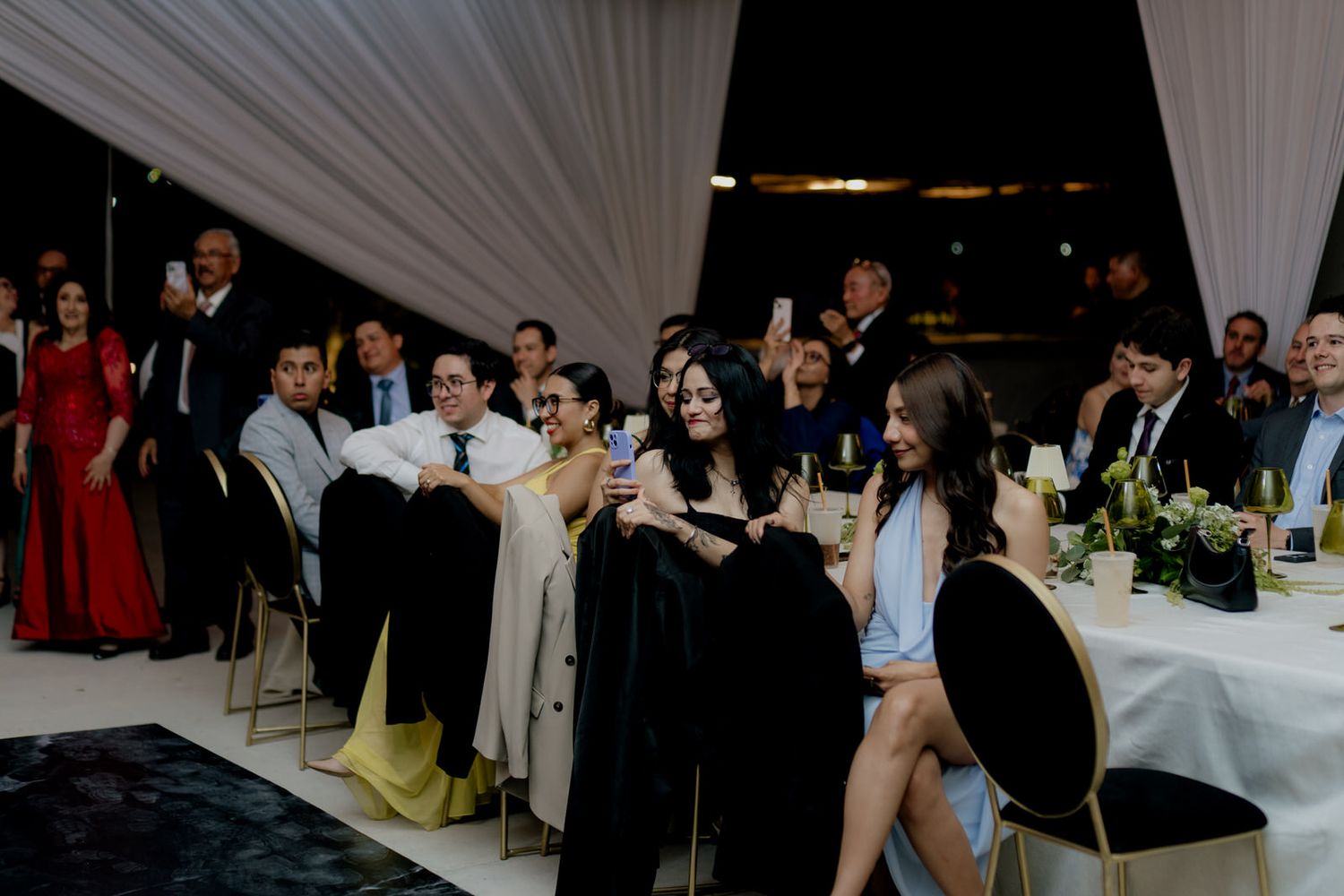 Wedding guests laugh and celebrate at elegantly decorated reception table under white draped tent during wedding party.