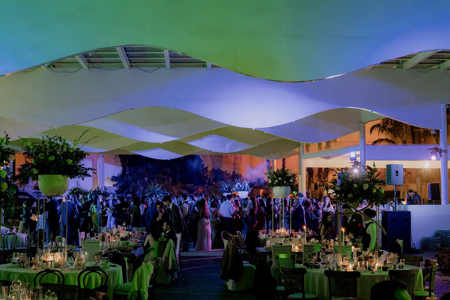 Elegant outdoor wedding reception with white wave canopy, green lighting, and guests dining at round tables.