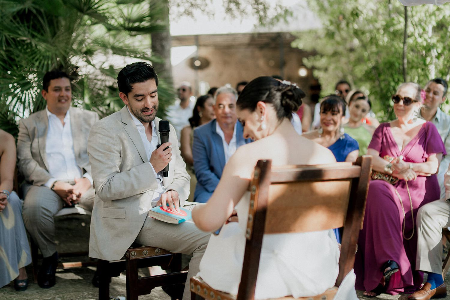 Guests celebrate at an intimate outdoor wedding reception under trees with natural lighting.