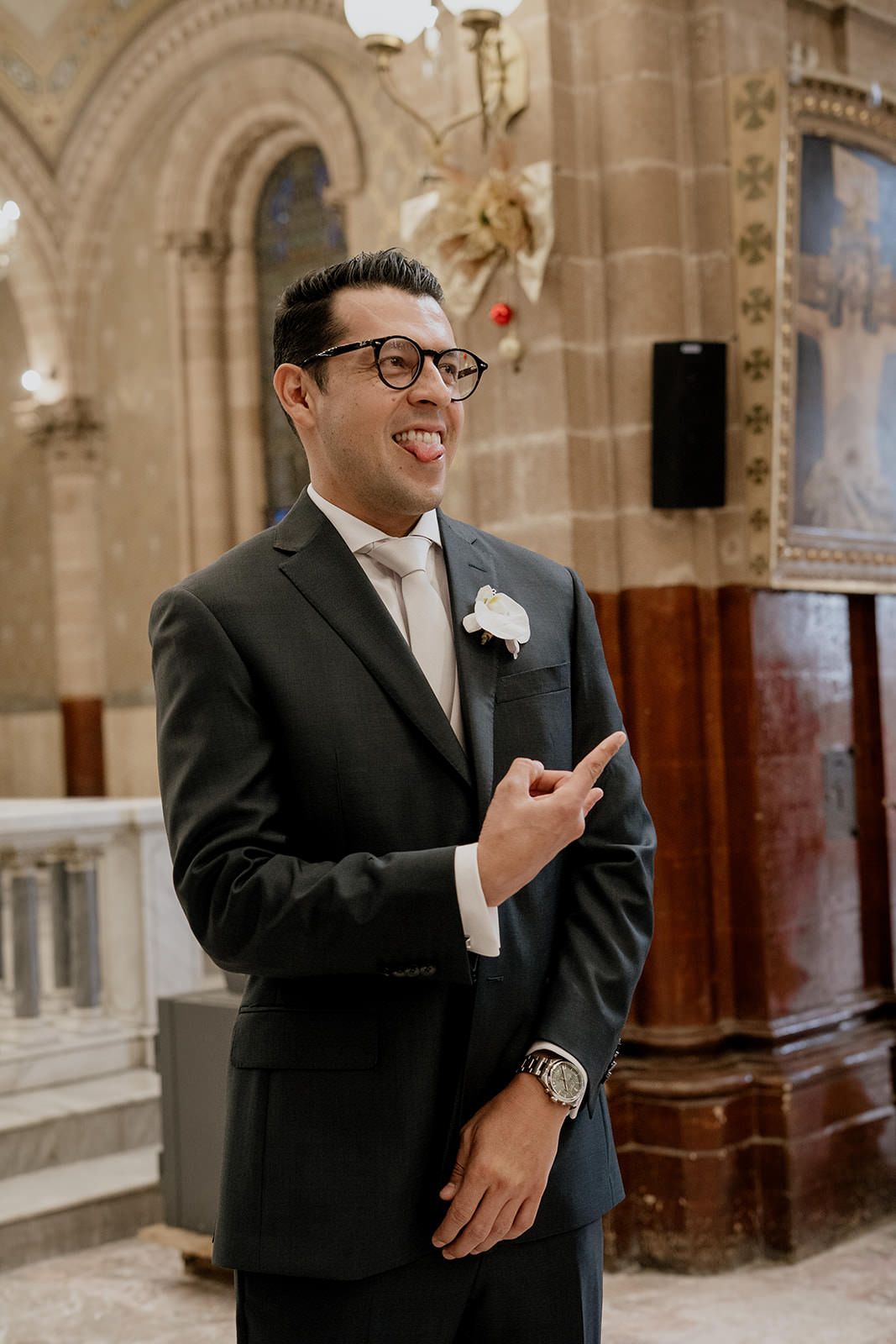 Groom in dark suit with boutonniere laughing and pointing inside an ornate church before wedding ceremony.