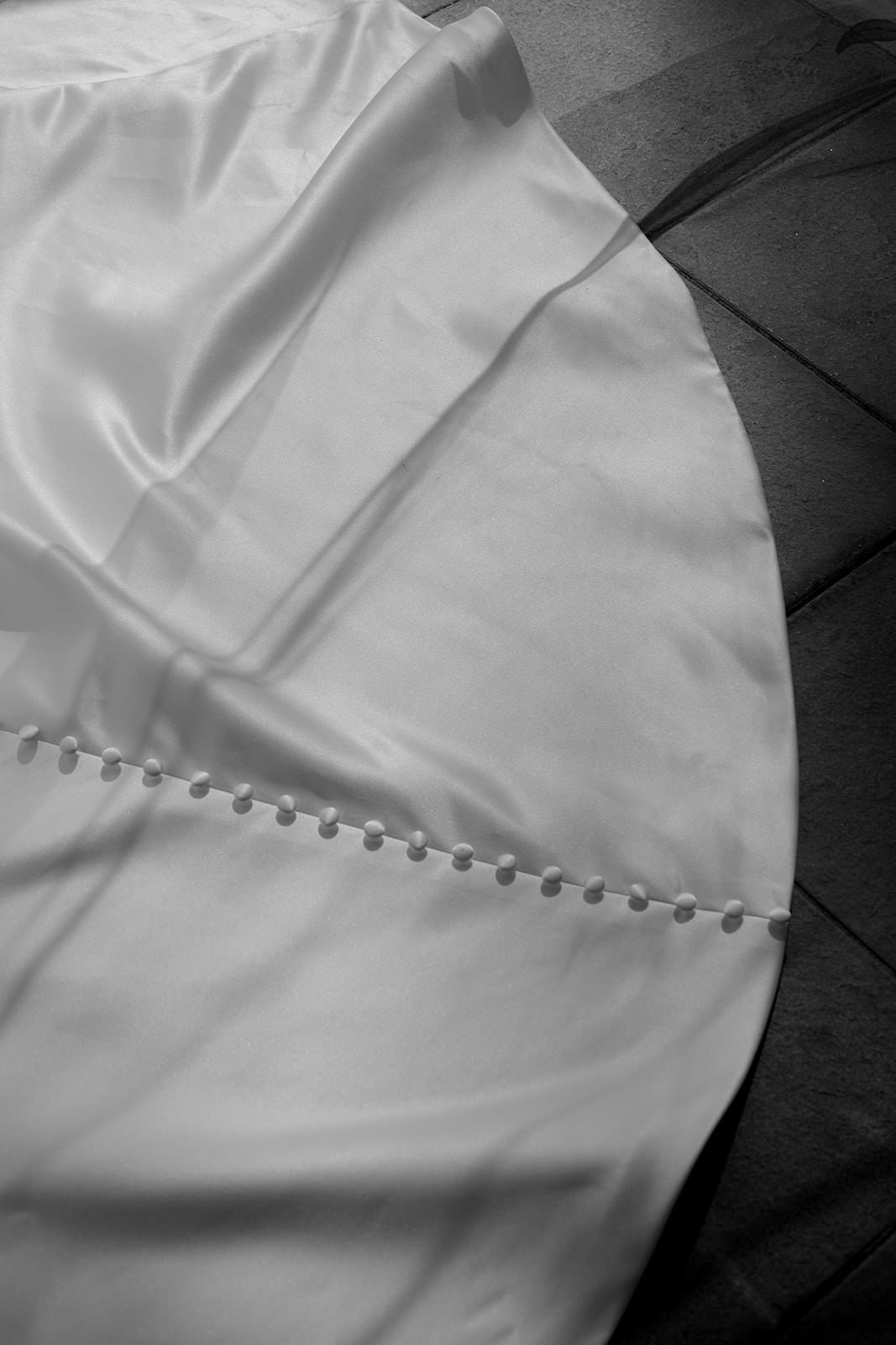Close-up black and white detail of elegant bridal gown hem with delicate pearl button trim on wooden floor.