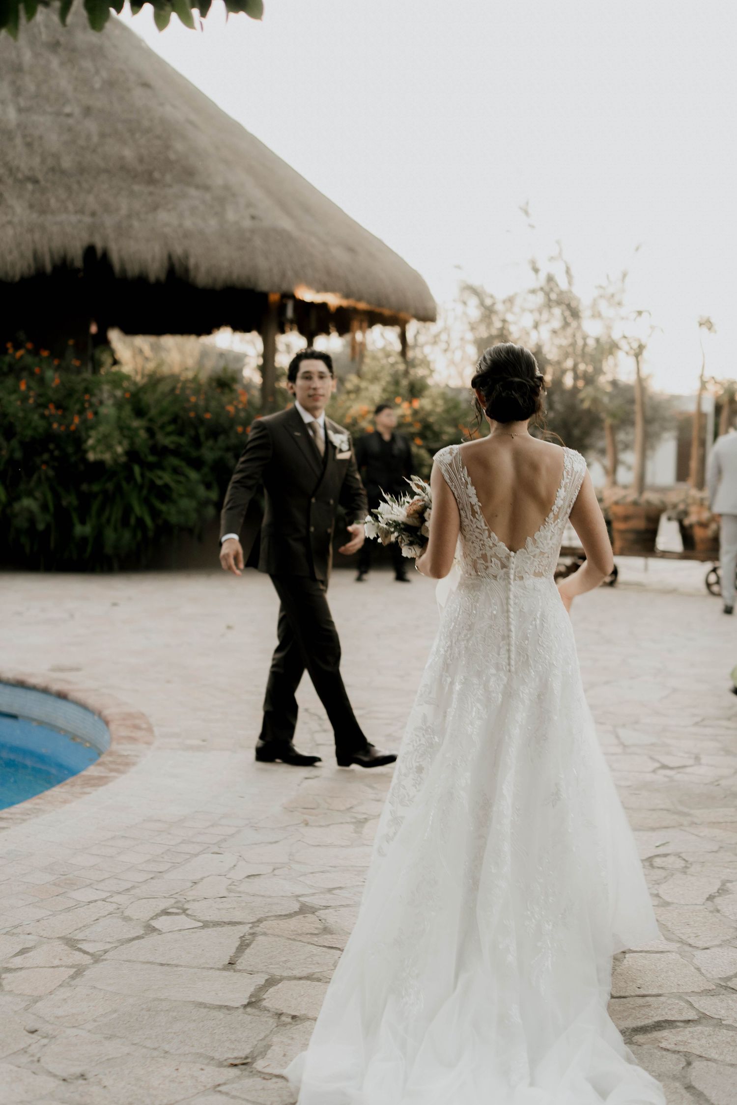 Couple in wedding attire walking along a stone path near a pool.