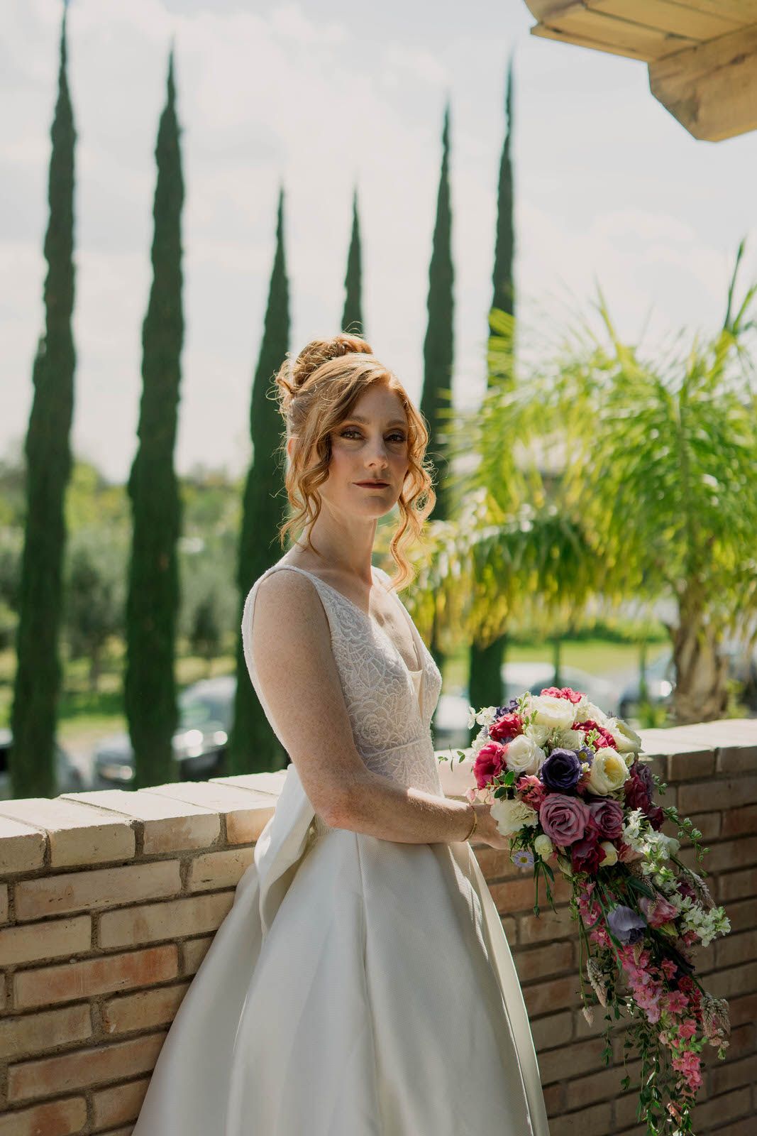 Elegant white wedding dress with flowing skirt and floral bouquet against a backdrop of cypress trees.