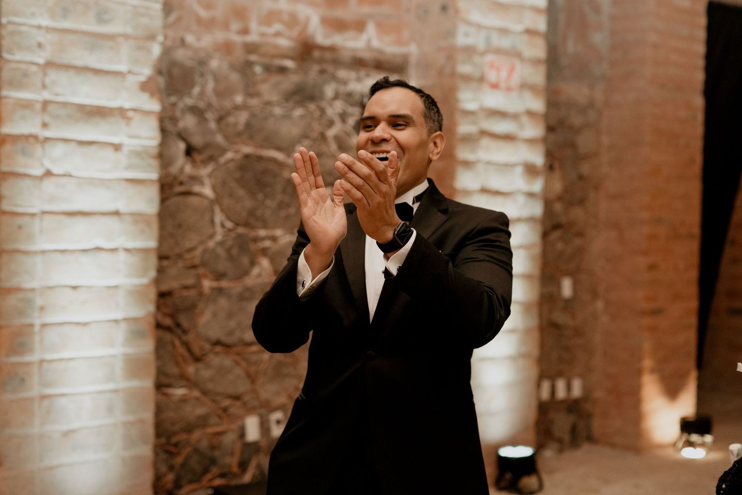 Wedding guests celebrate in an illuminated brick venue with hands raised in celebration.
